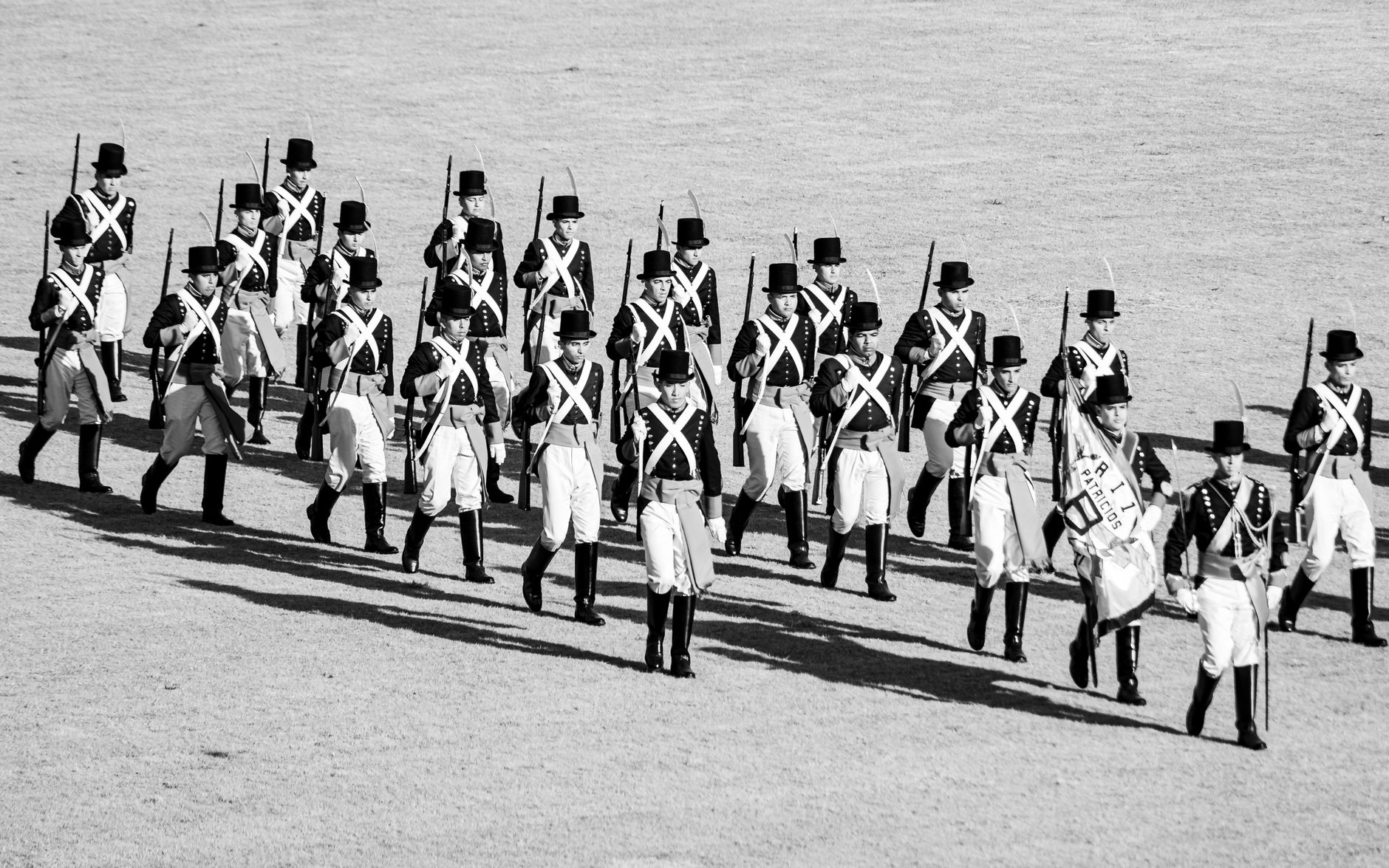 A black and white image of soldiers in historical military uniforms marching on a field.