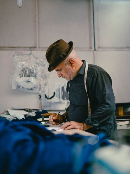 Free Elderly tailor working attentively at a sewing table with measuring tape around his neck in a workshop setting. Stock Photo