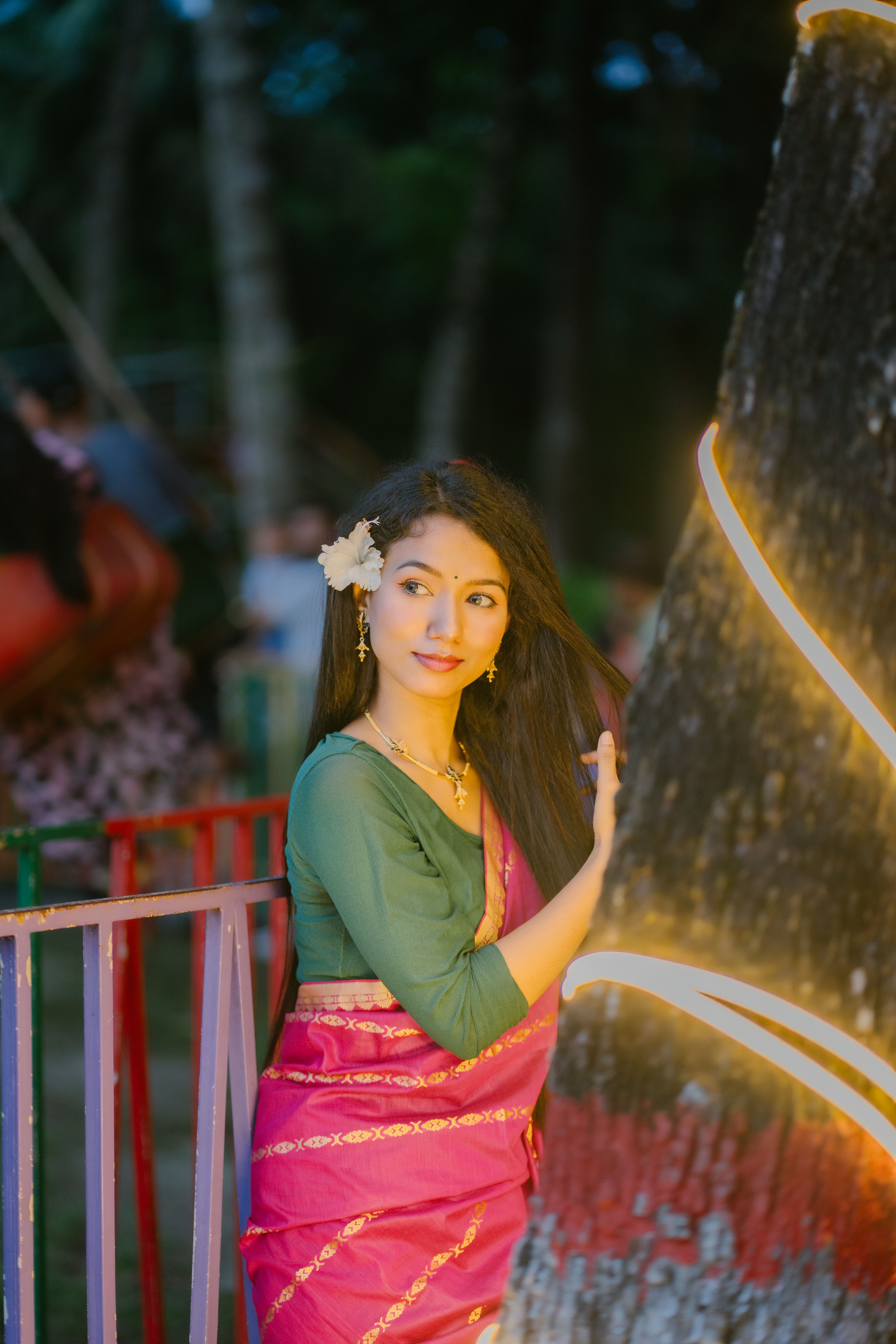 A young woman in a pink saree poses gracefully beside a lit tree during a festive evening.