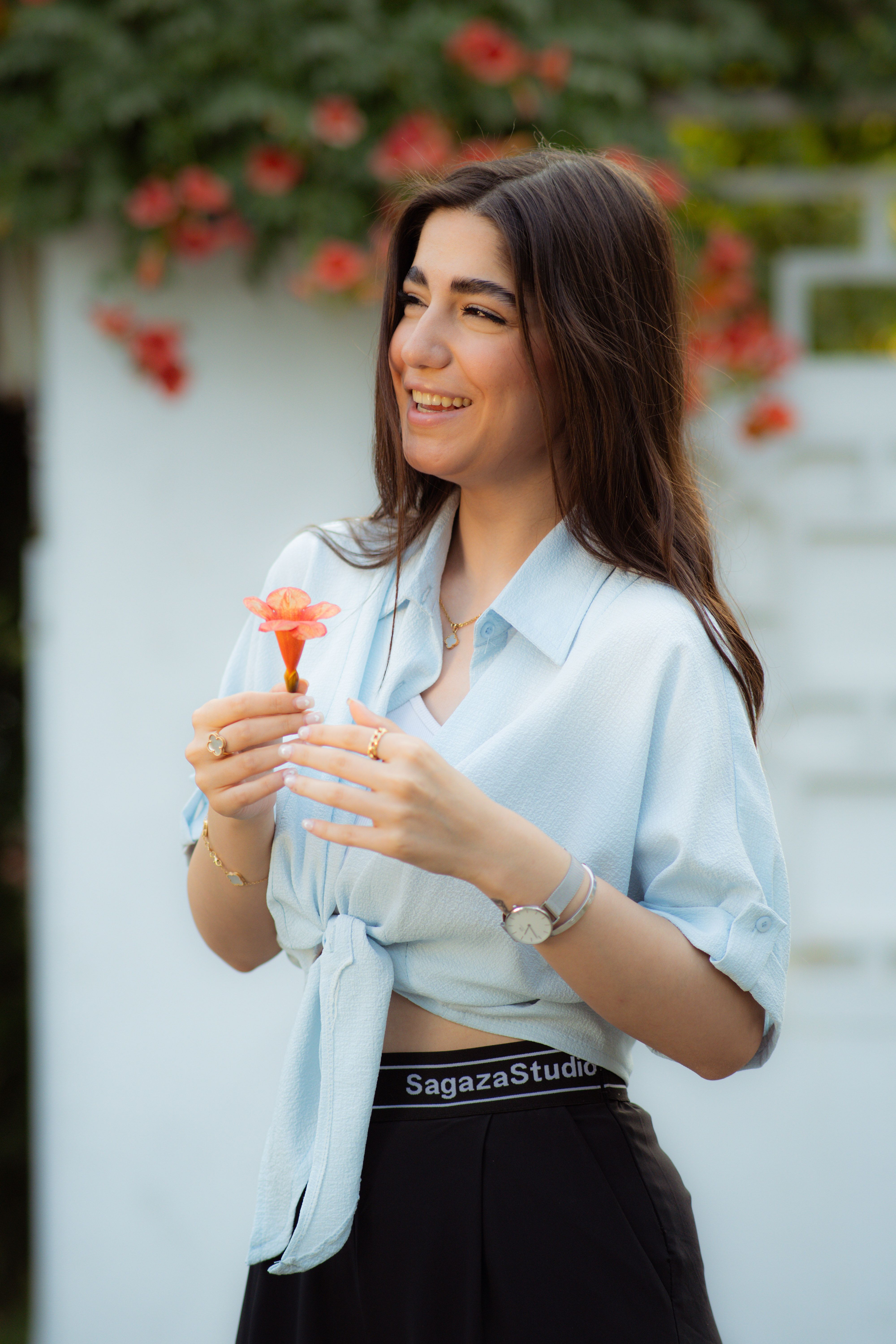 A joyful woman in a light blue shirt holds a flower, standing outdoors with a floral background.