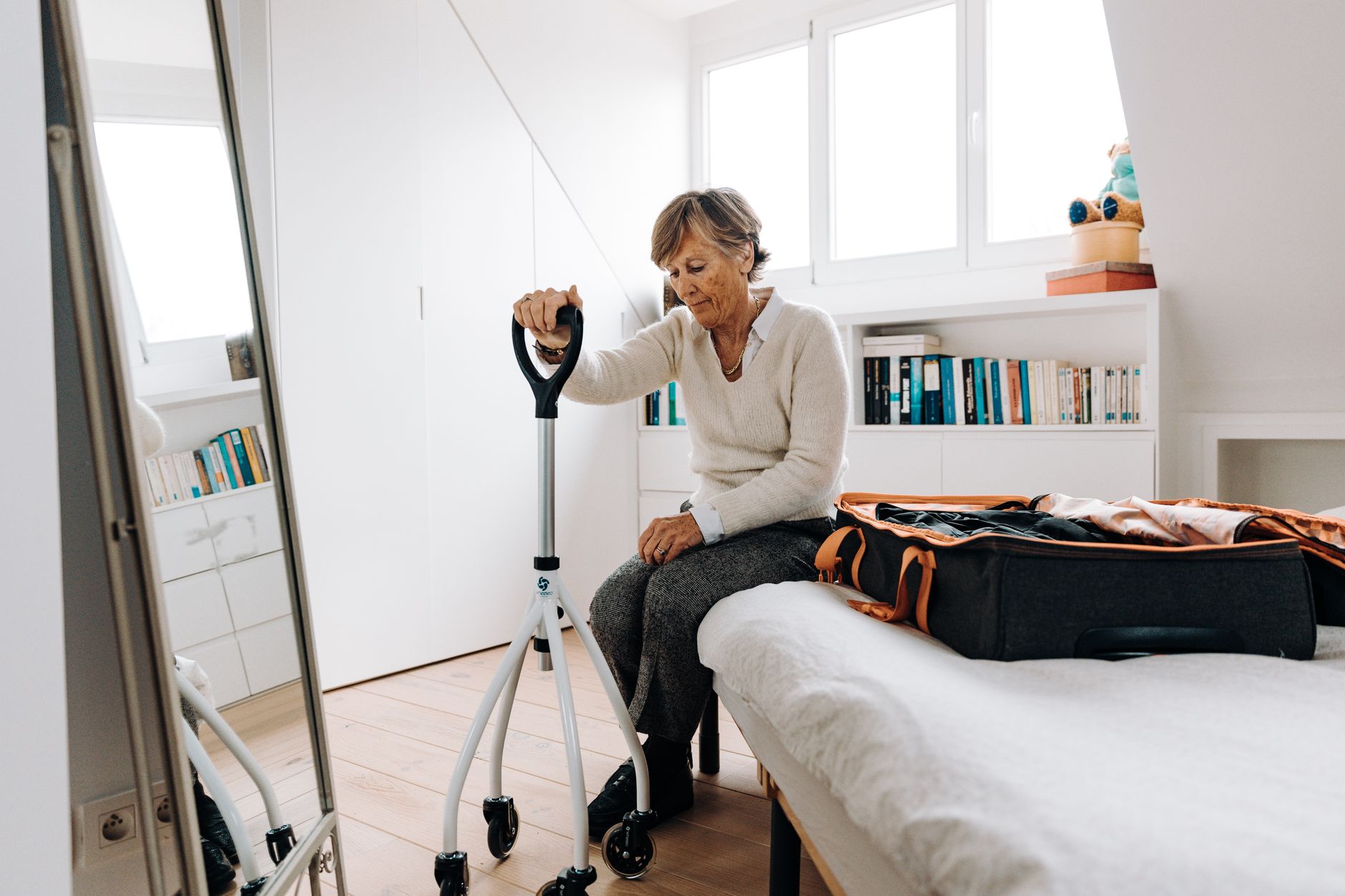 Senior woman sitting on bed with walker, preparing suitcase in bright bedroom.