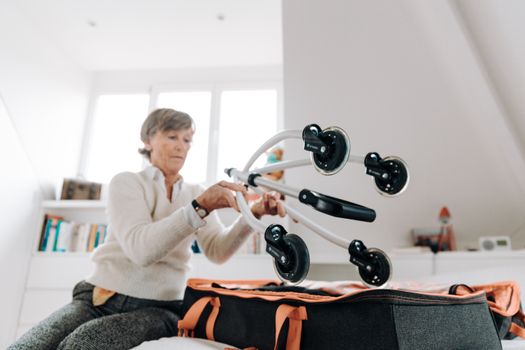 Elderly woman packing a rollator in a suitcase in a bright room.