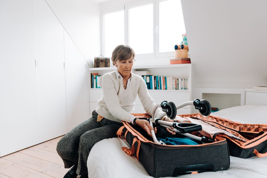 Senior woman packing a rollator in a suitcase in a cozy bedroom setting. Bright indoor lighting.