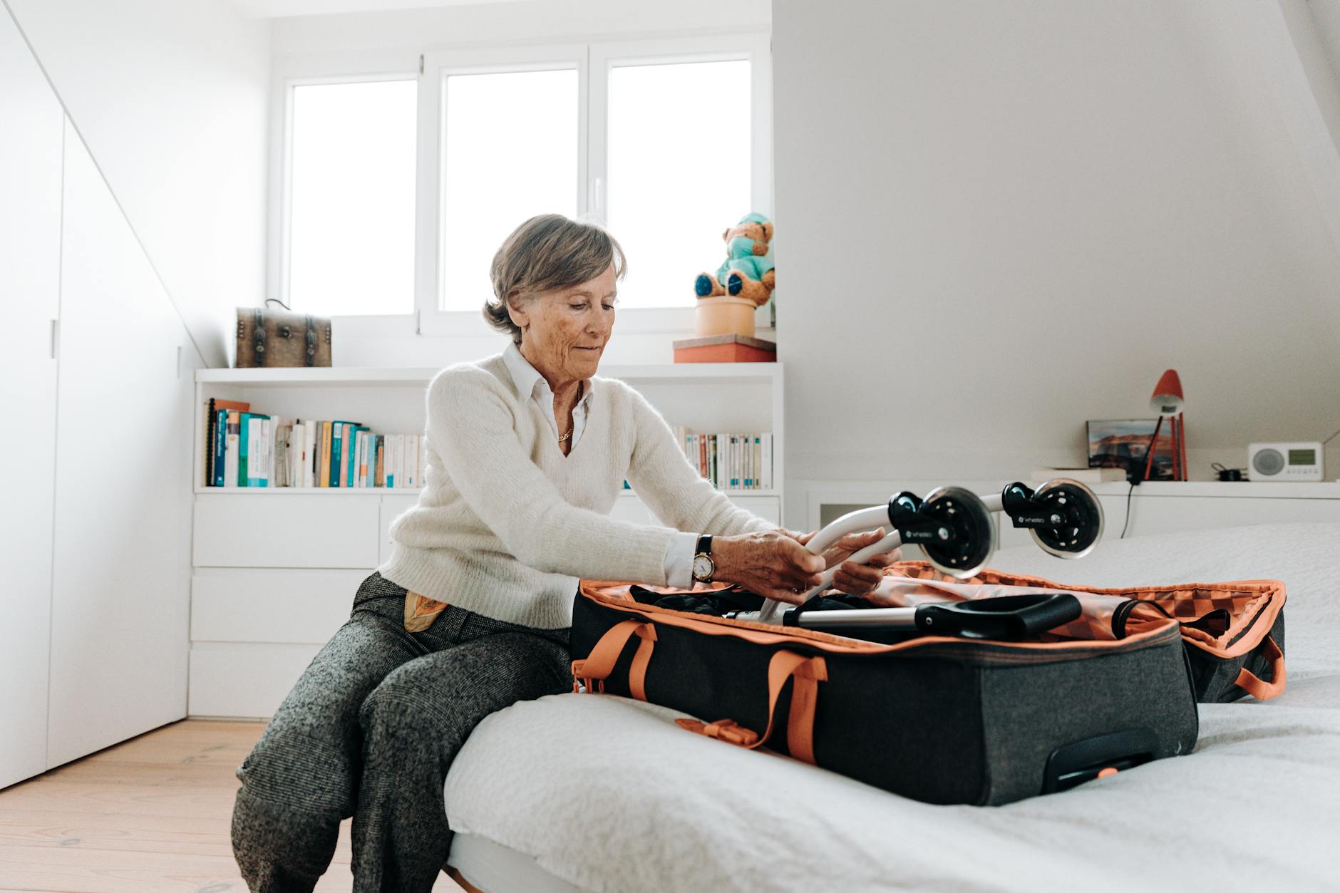Elderly woman packing a rollator into a suitcase at home, preparing for a trip.