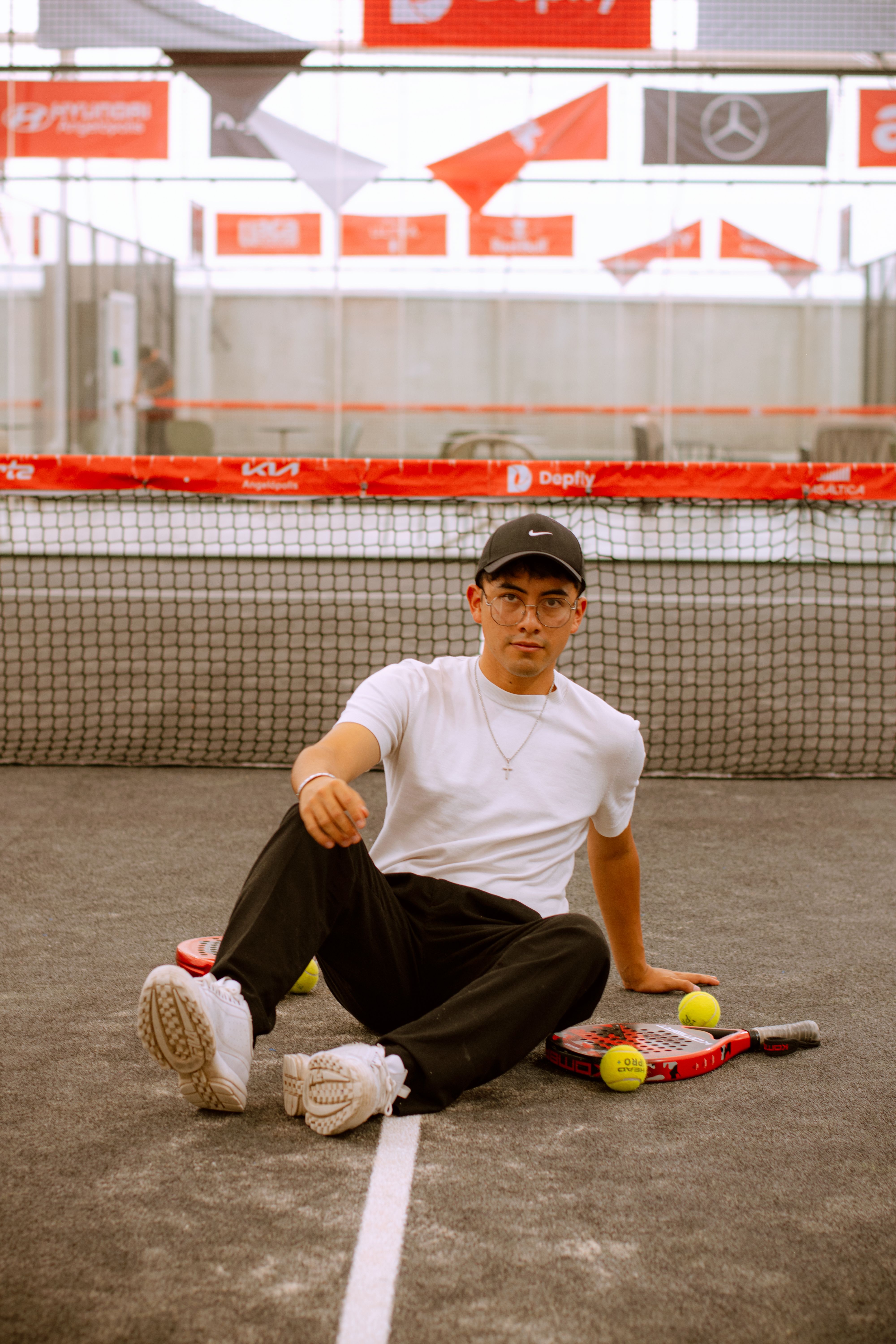 A young man sits on a padel court with rackets and tennis balls.