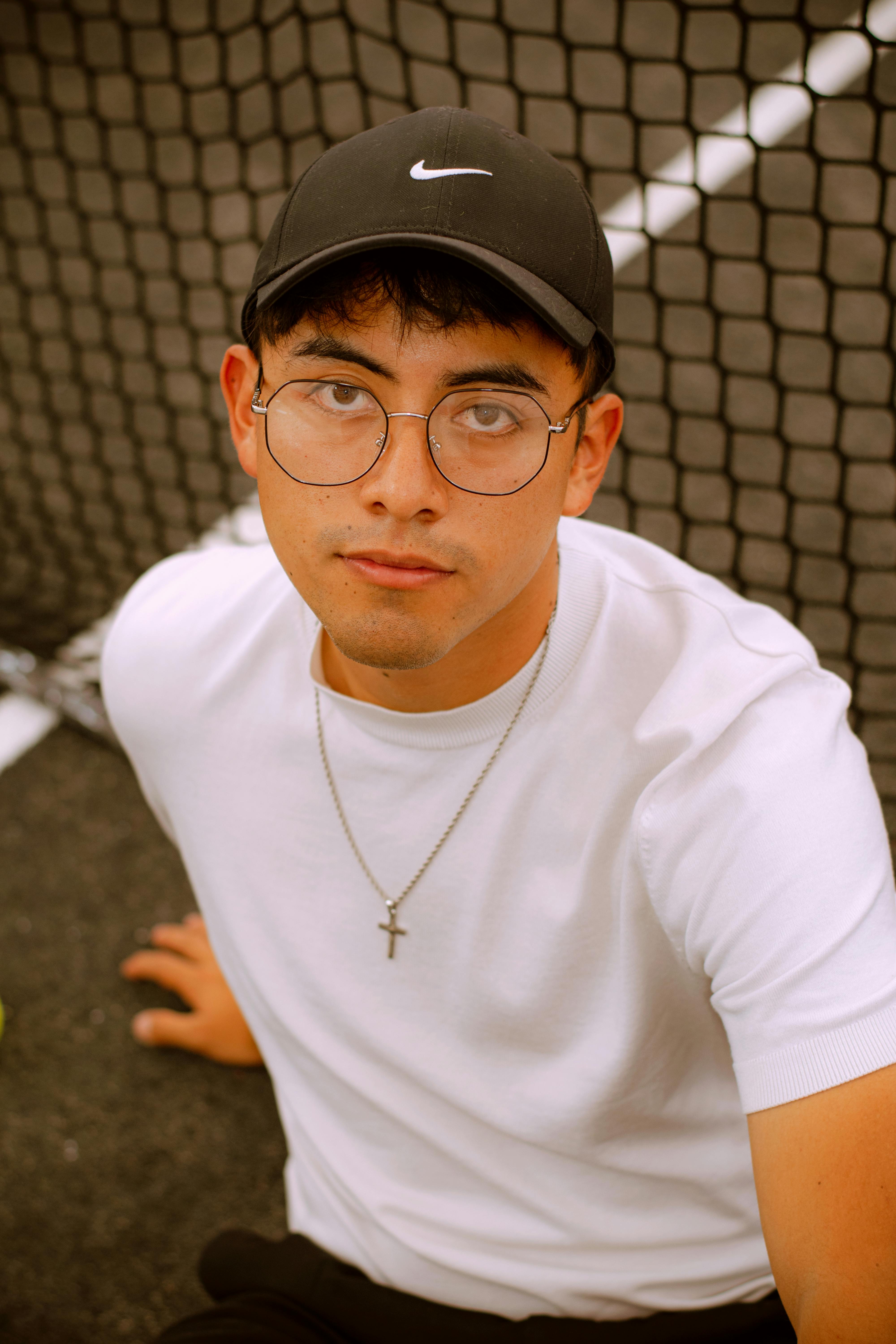 Portrait of a young man in casual attire and glasses sitting by a tennis court net.