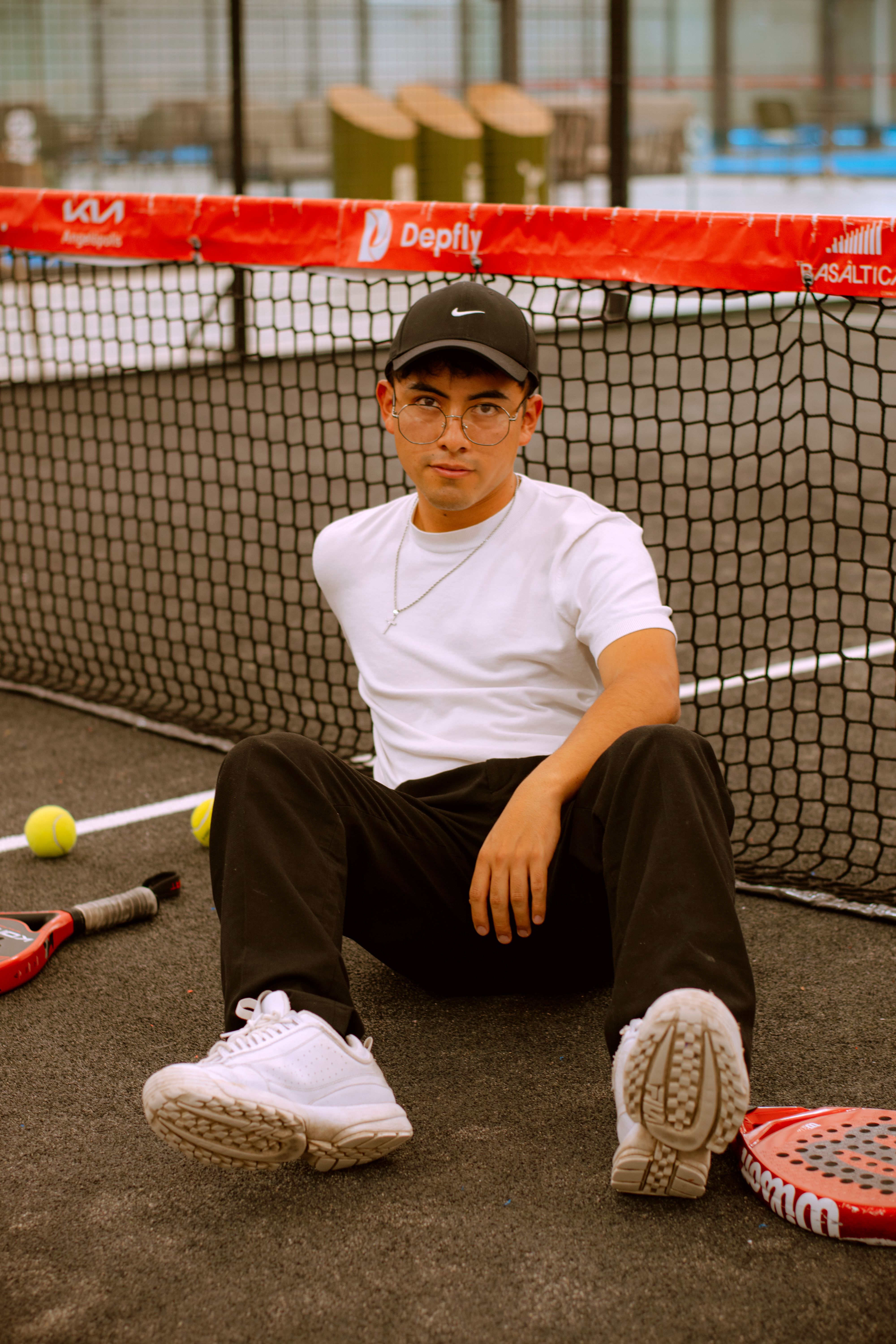 A young man sits casually on a padel court with equipment by his side, exuding a sporty and relaxed vibe.