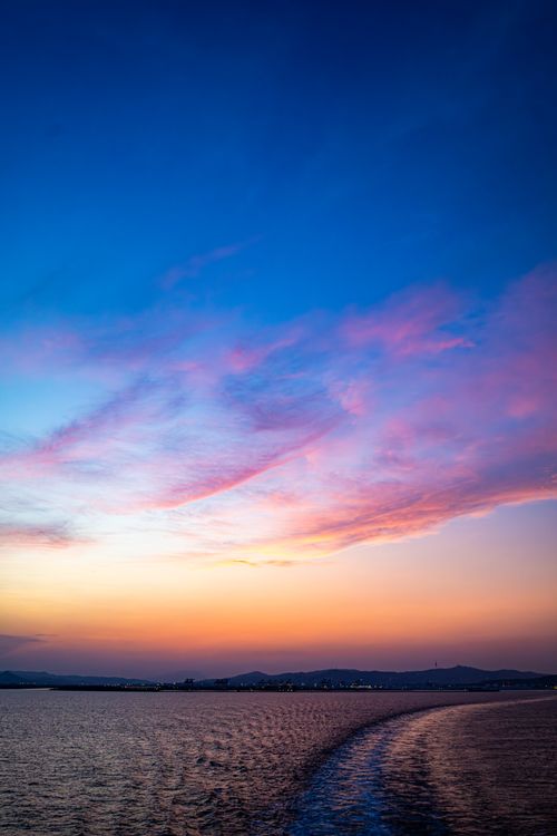 Free A breathtaking sunset over ocean waters with a city's twinkling lights under vibrant, colorful skies. Stock Photo