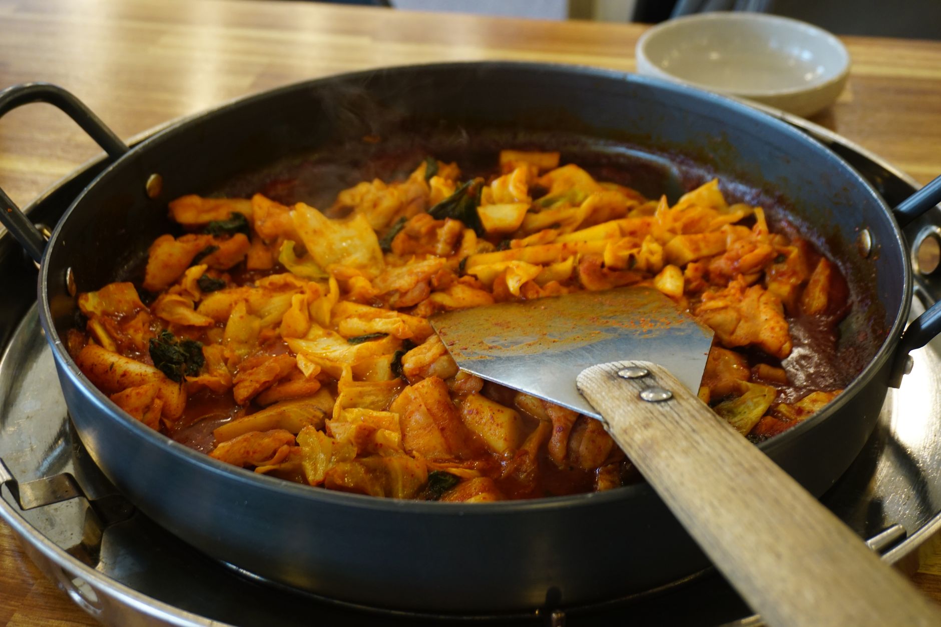 A skillet of Korean dak galbi with golden chicken pieces, colorful vegetables, and rice cakes coated in glossy red-brown gochujang sauce, garnished with scallions and sesame seeds