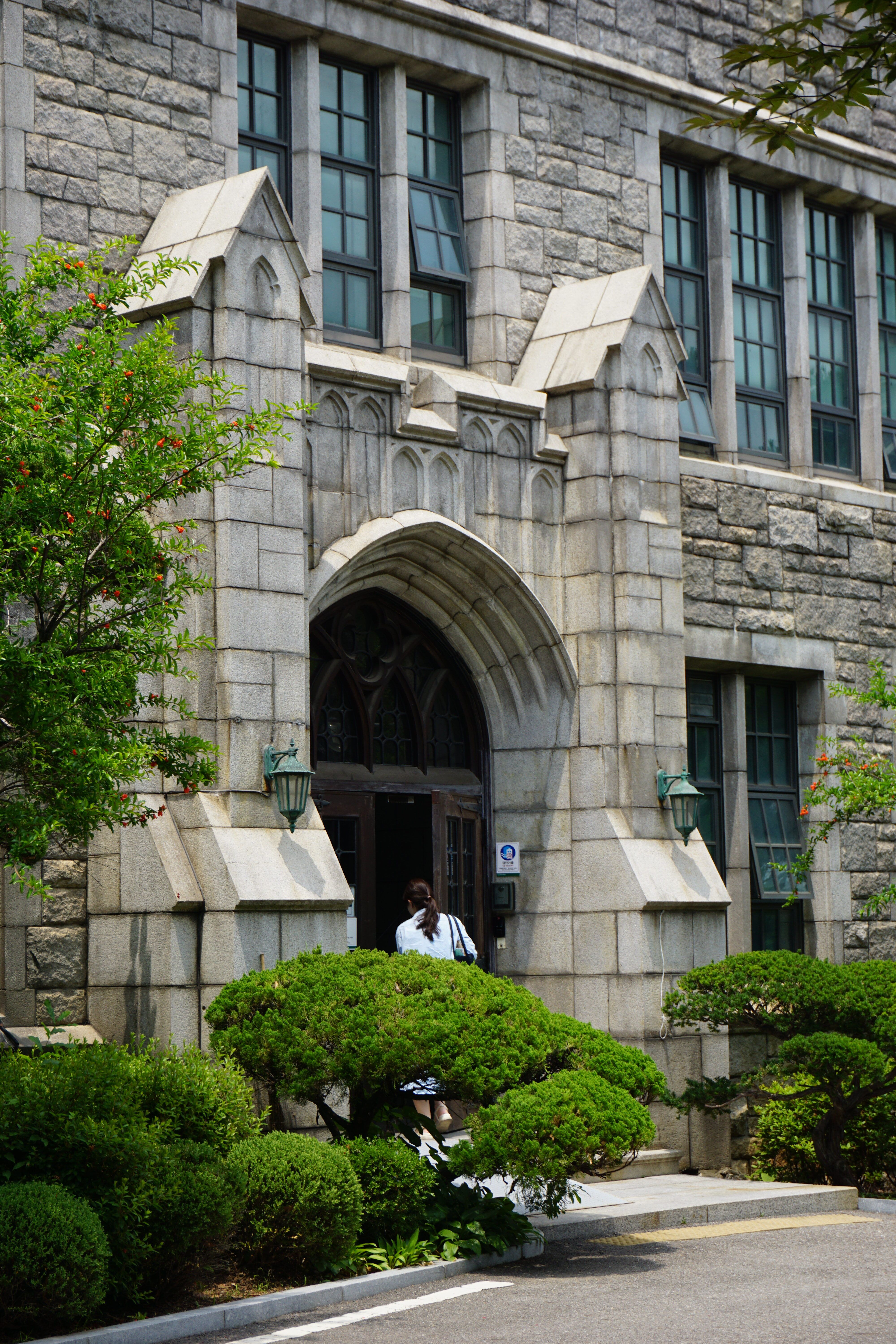 A person stands in front of a historic stone building with Gothic arches and lush greenery.