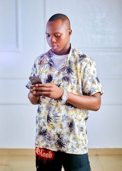 A young adult indoors engaging with a smartphone, wearing a floral shirt and casual pants.