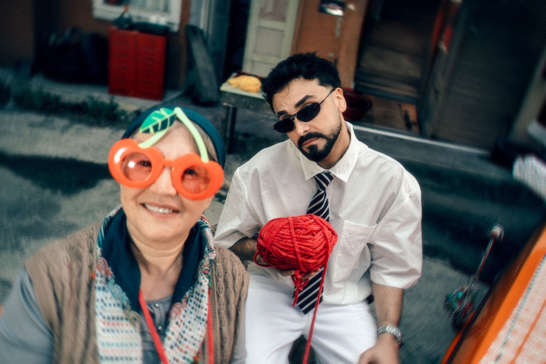 Outdoor portrait of two adults with quirky glasses and a red yarn, set in a lively urban area.