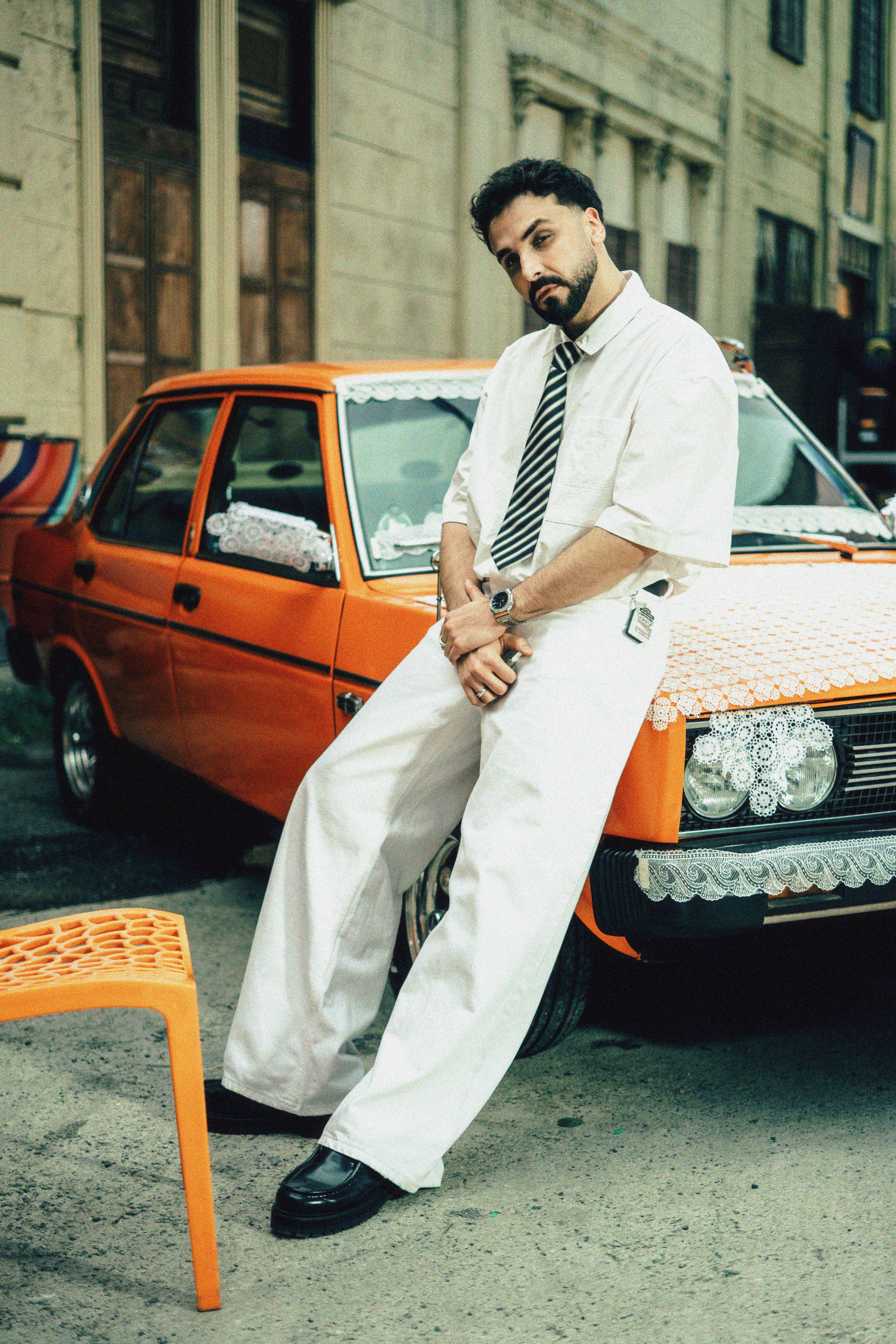 A man wearing white clothes and a tie leans against an orange car in an urban setting.
