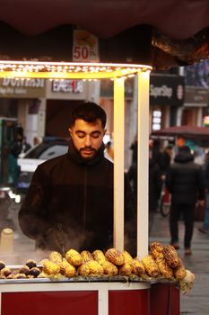 A street vendor sells roasted corn on a bustling city street at night under warm lights.