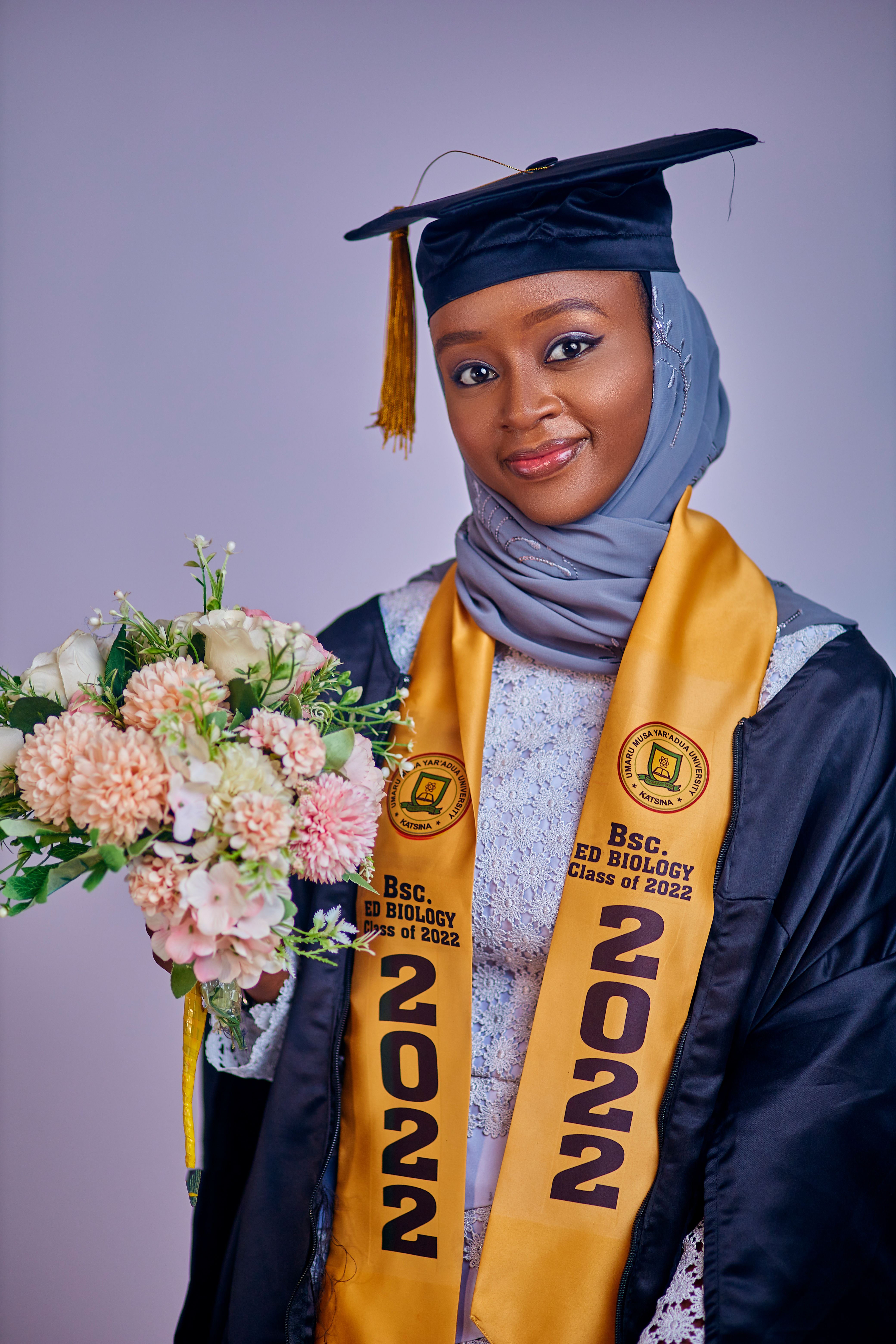 Smiling young graduate in cap and gown holding a bouquet of flowers. Class of 2022.