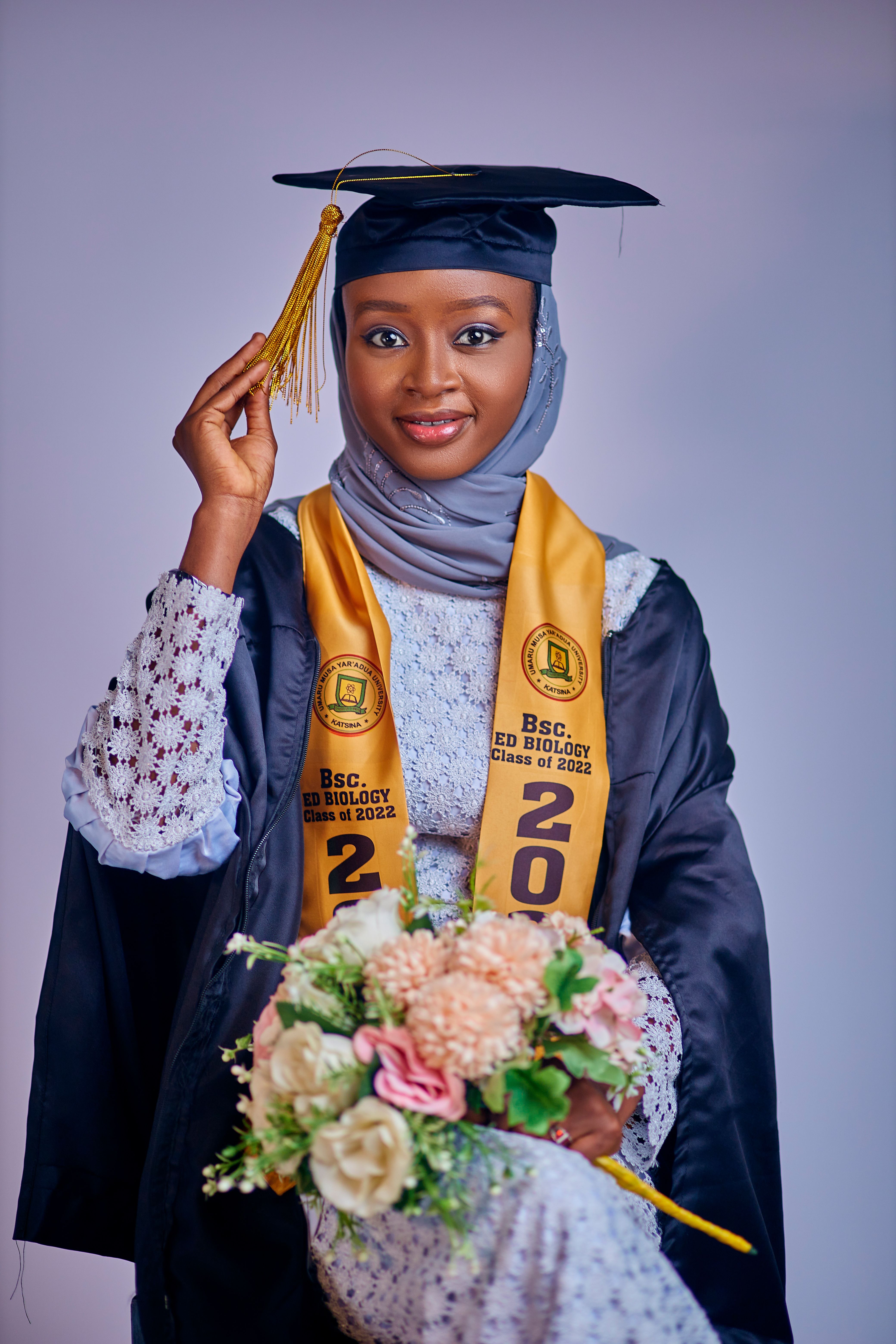 Young woman in graduation cap and gown, holding flowers, smiling at the camera.