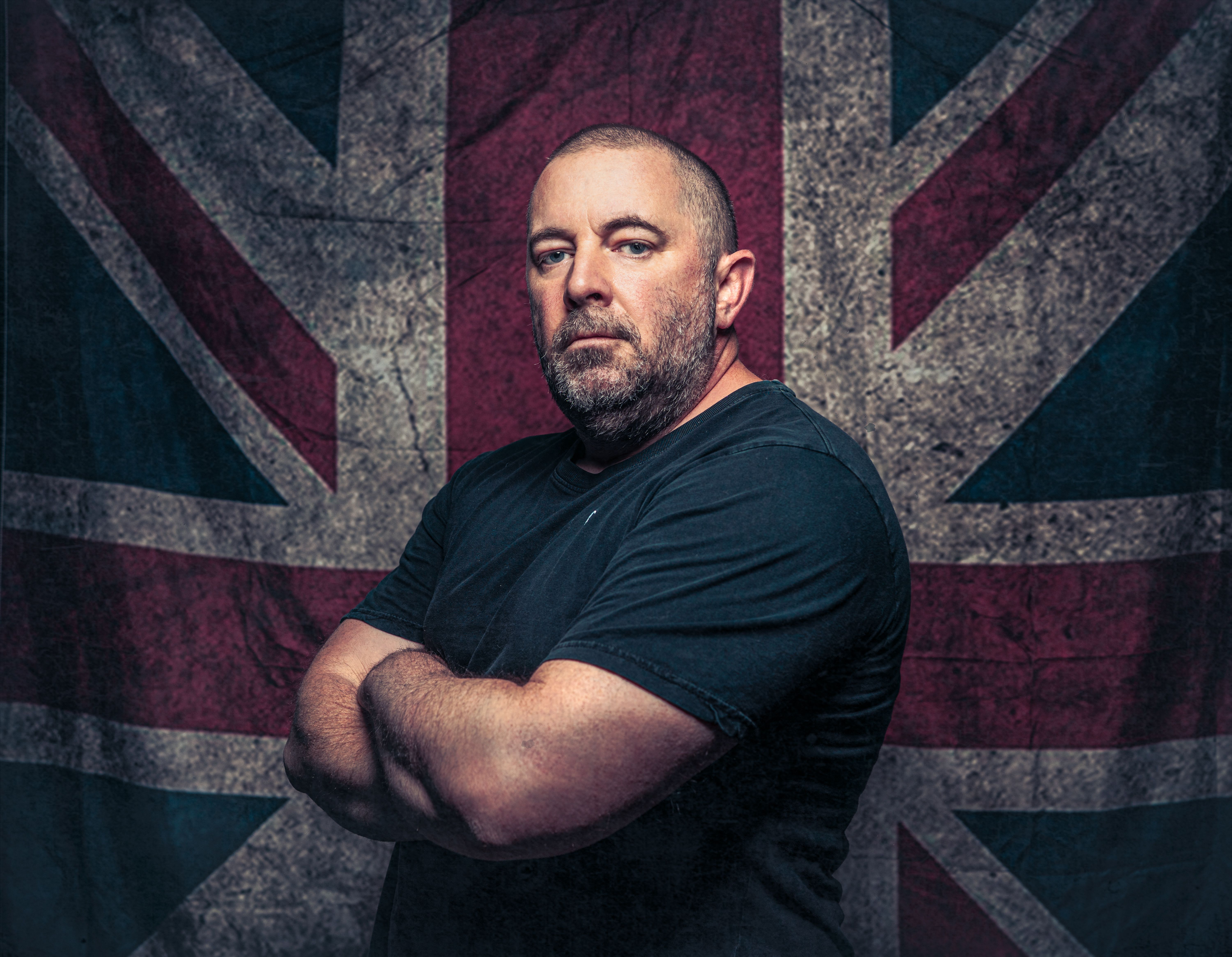 A confident man poses with arms crossed against a rustic British flag backdrop, showcasing strength and patriotism.
