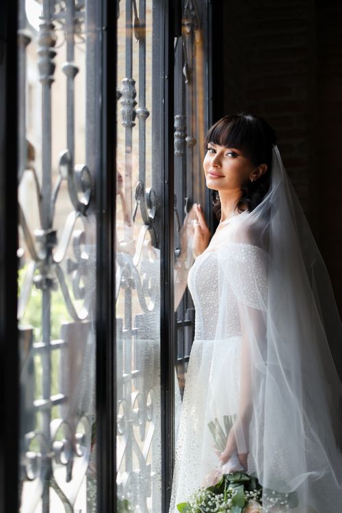 Free Beautiful bride in wedding gown gazing out the window, holding white flowers. Indoors setting with soft natural light. Stock Photo