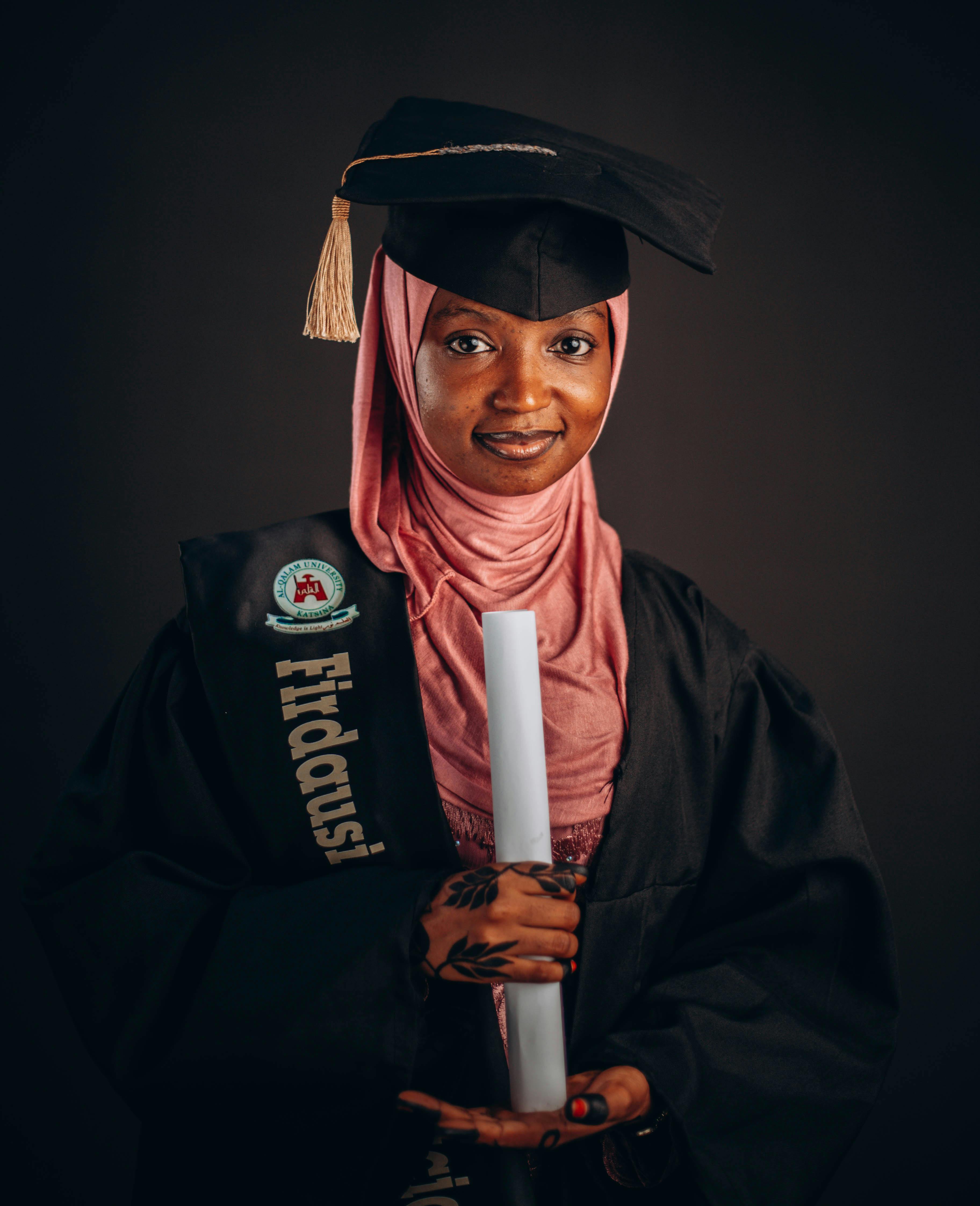 Confident woman wearing graduation gown and hijab holding diploma, celebrating academic achievement.