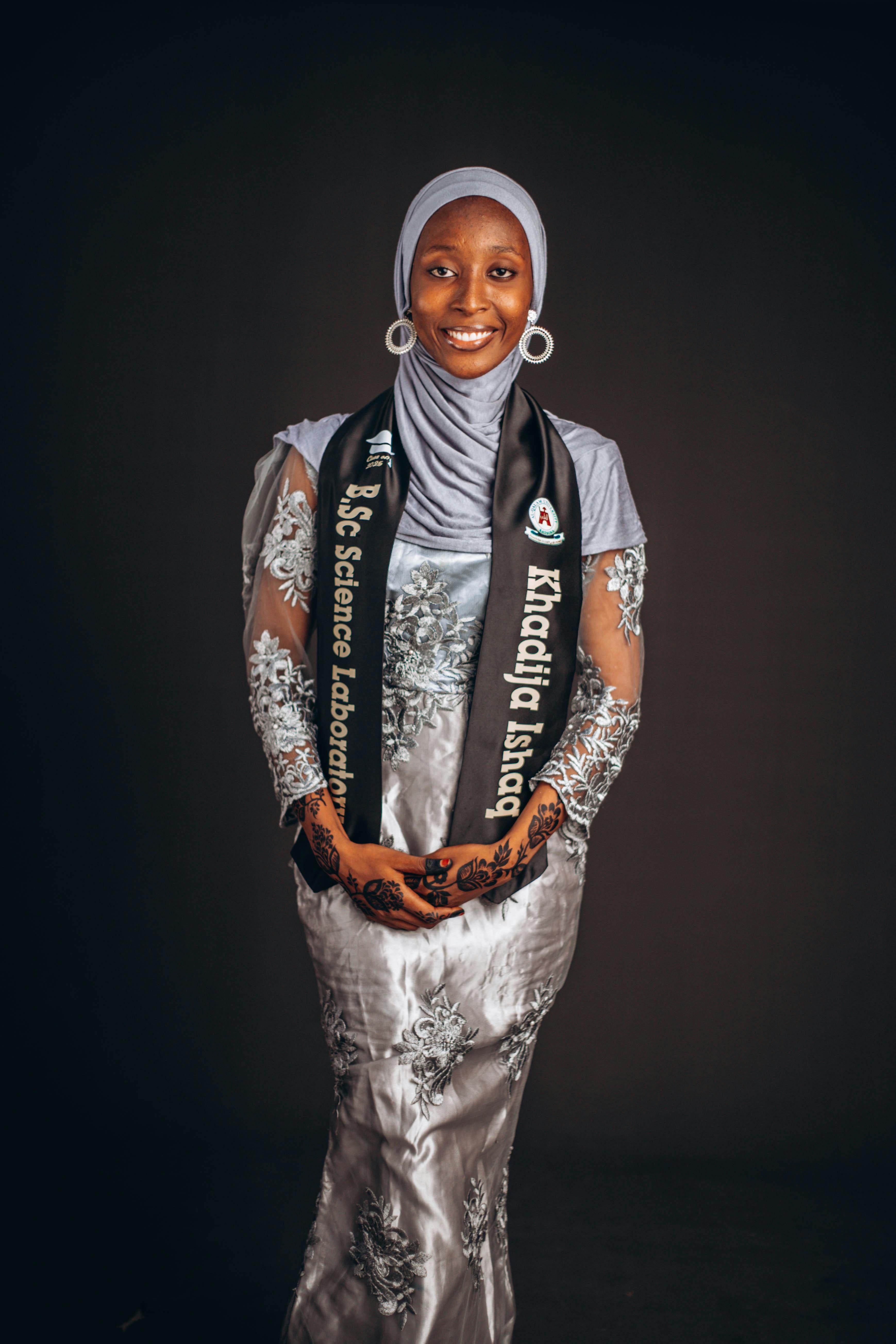 A smiling woman in a hijab and graduation stole, posing with confidence.
