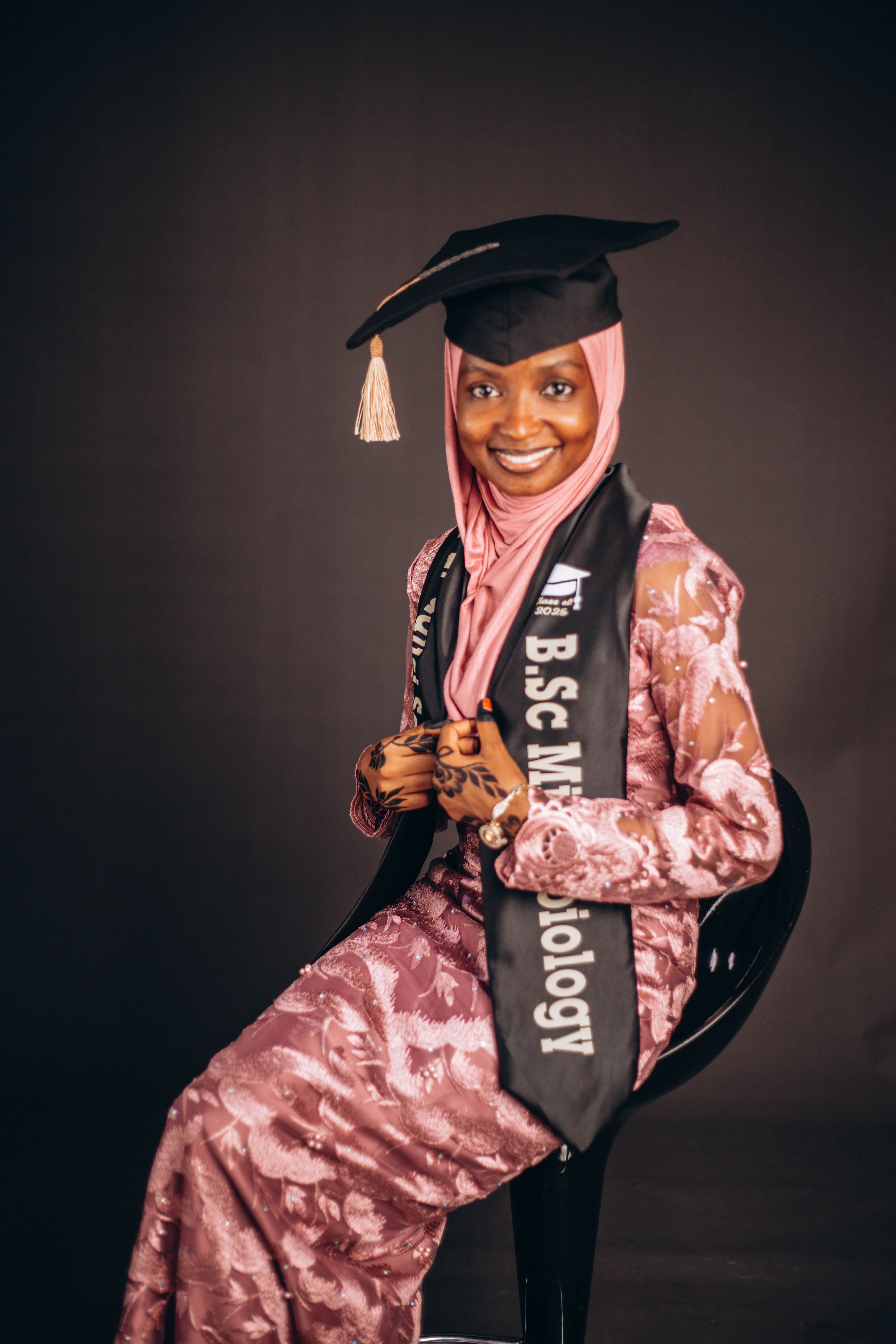 A young woman celebrating her graduation in a pink dress and hijab, holding a microbiology degree.