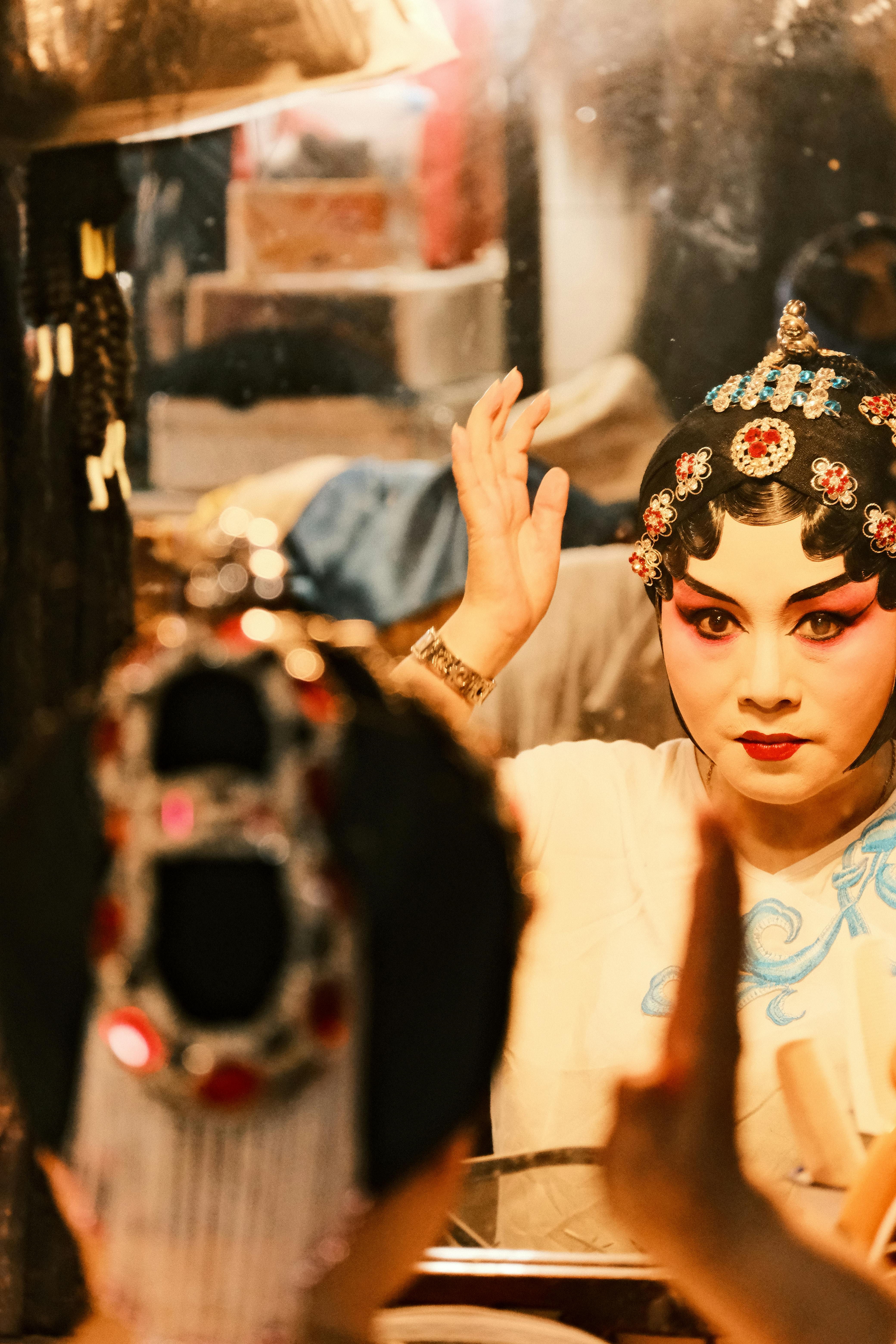 Gratis Un'artista dell'opera cinese con un trucco colorato si sistema il copricapo davanti allo specchio. Foto a disposizione