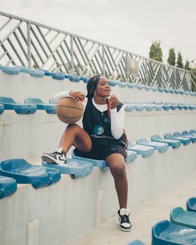 Confident young athlete holding basketball seated in outdoor stadium bleachers.