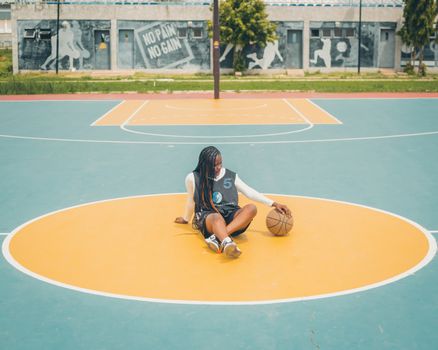 Young woman in basketball jersey sitting on an outdoor court with a ball.