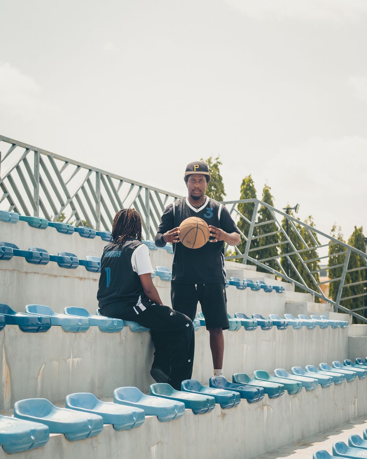 Two Black athletes in athletic gear in a stadium-stands setting; one stands holding a basketball, the other is seated in a jersey, both mid-conversation in a coaching moment.