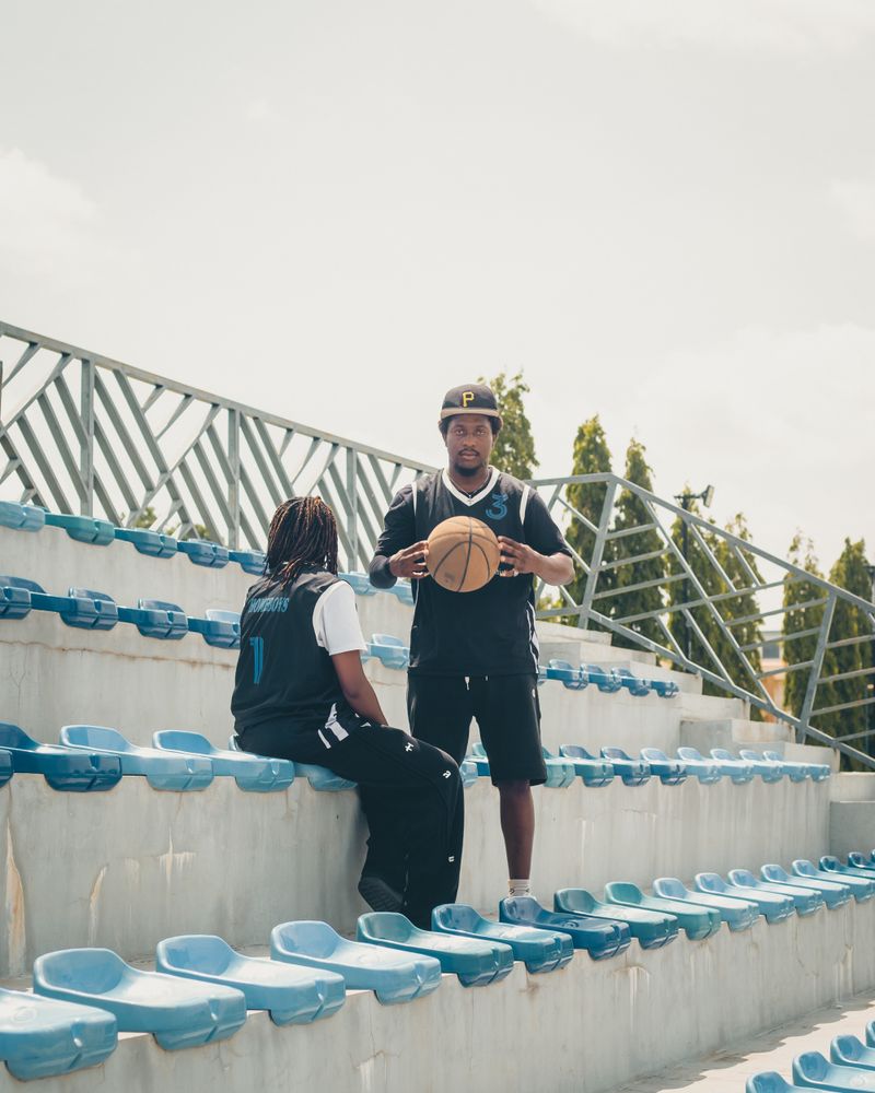 Two Black athletes in athletic gear in a stadium-stands setting; one stands holding a basketball, the other is seated in a jersey, both mid-conversation in a coaching moment.