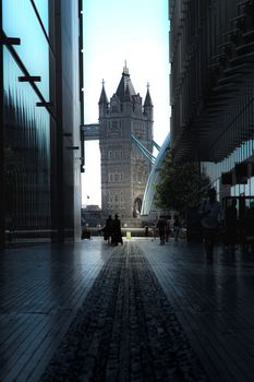 Silhouette view of people walking towards iconic Tower Bridge in London through a modern alleyway.
