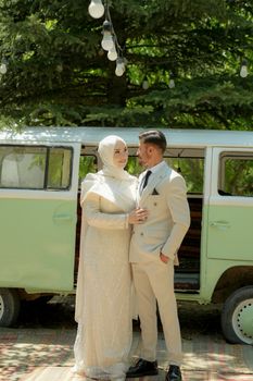 Stylish couple in formal attire standing beside a vintage mint green van surrounded by greenery.