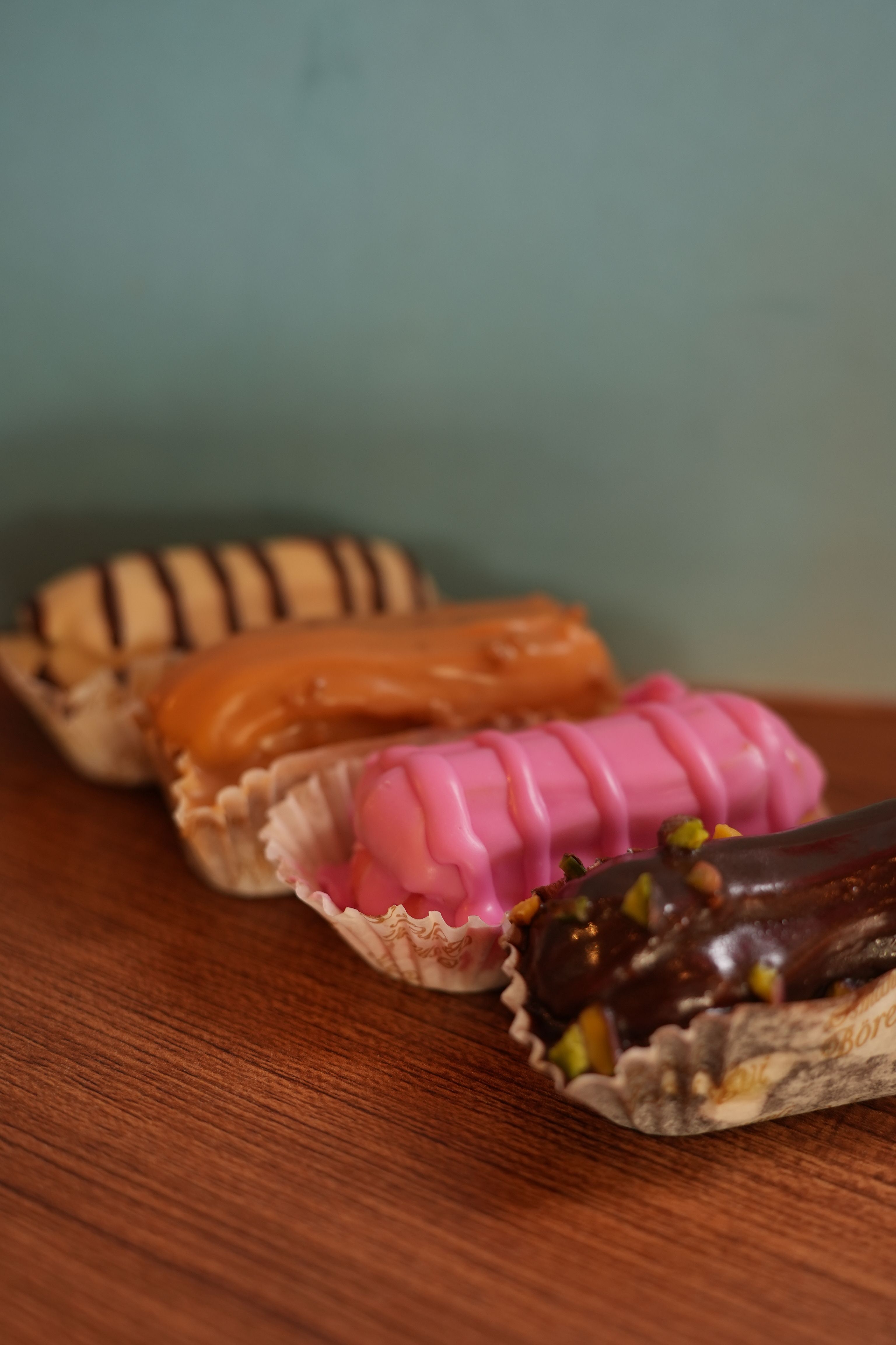 Four vibrant eclairs in a row on a wooden surface with a pastel backdrop, offering a delectable treat for dessert lovers.