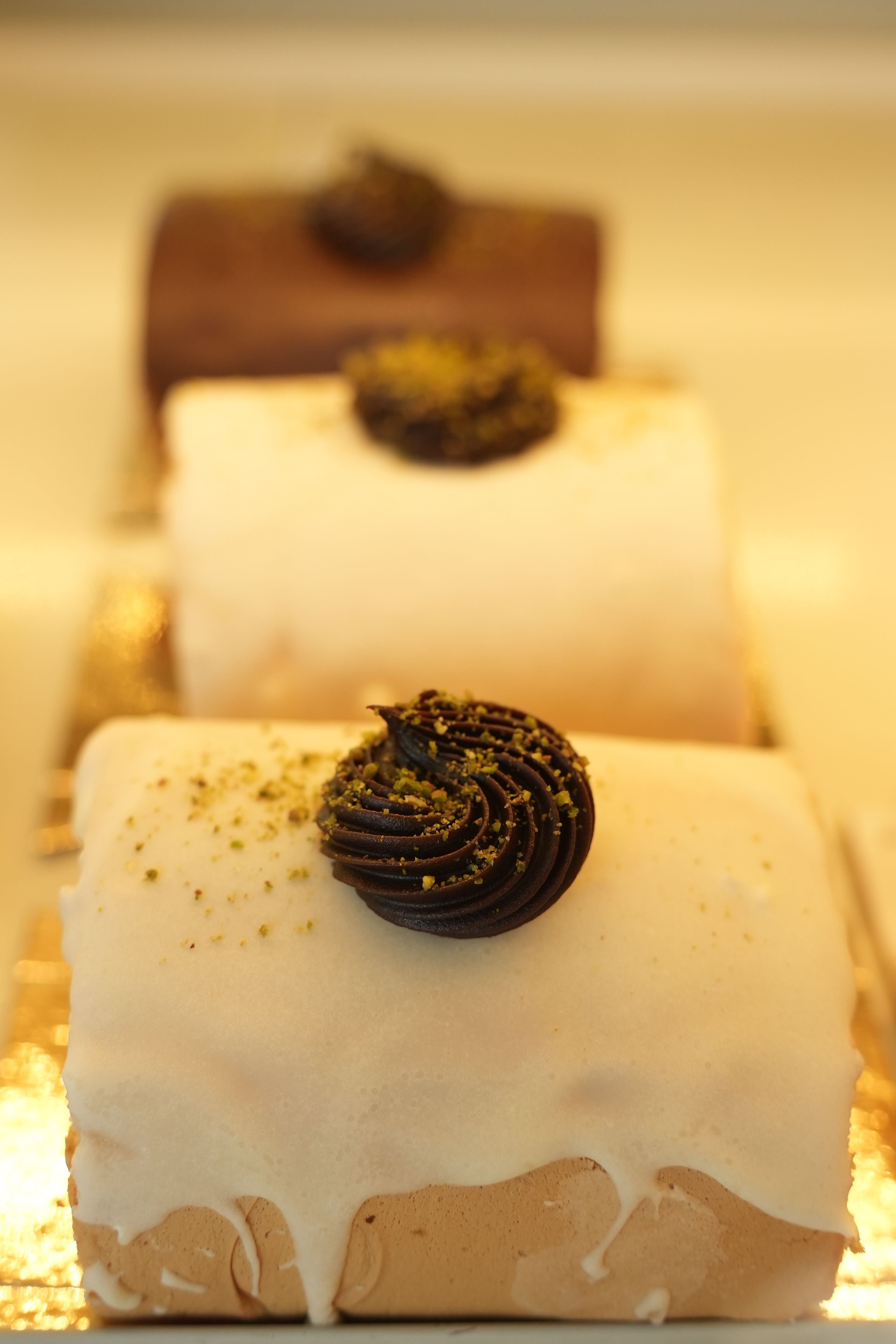 Assorted frosted cakes topped with chocolate and pistachio in a bakery.