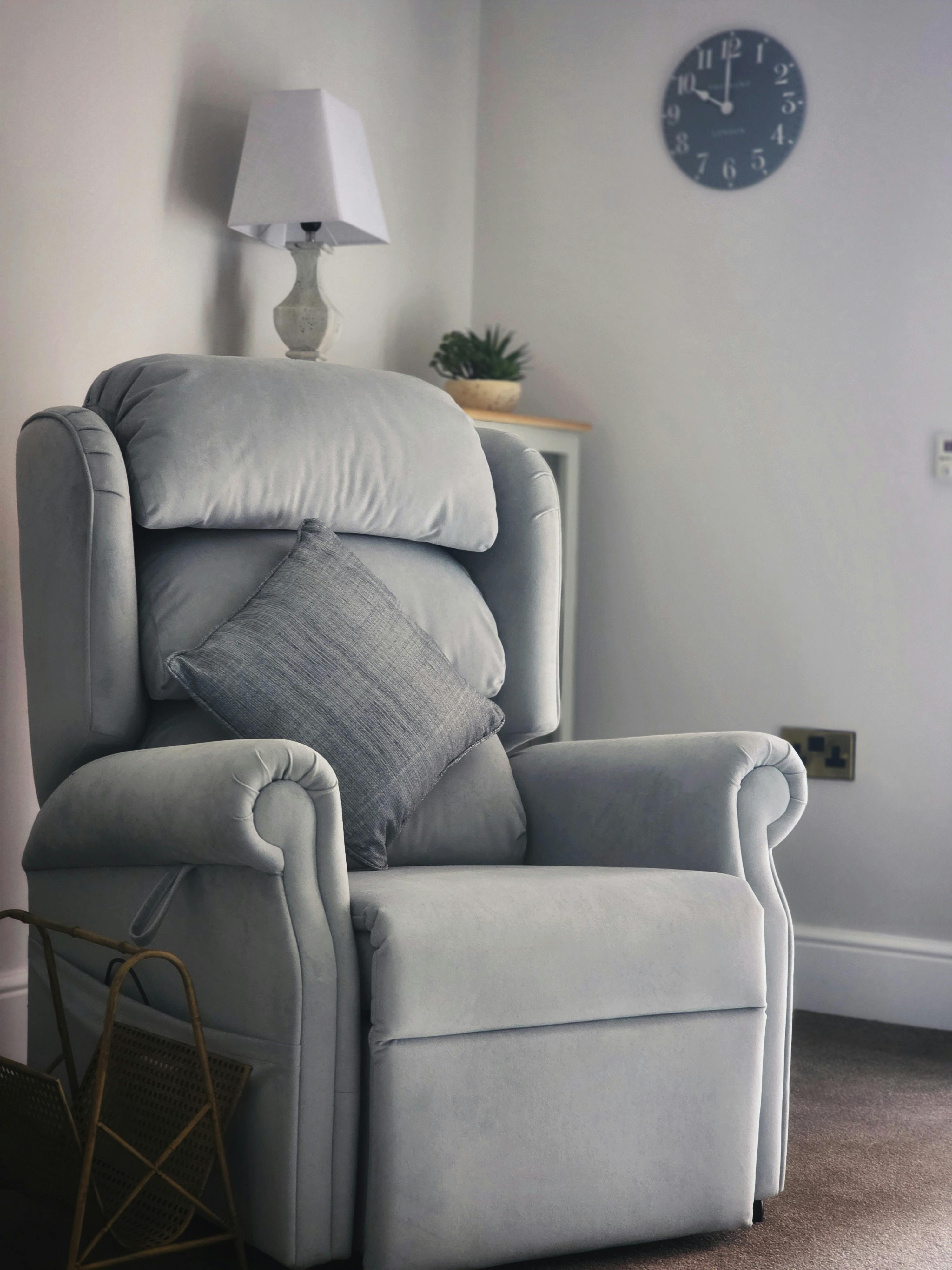 A plush grey armchair with pillow in a serene living room corner, featuring a clock and lamp.
