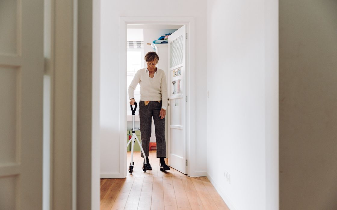 Older woman walking down a bright hallway at home with the support of a rollator