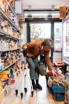A senior woman with a rollator shopping for produce in a grocery store aisle.