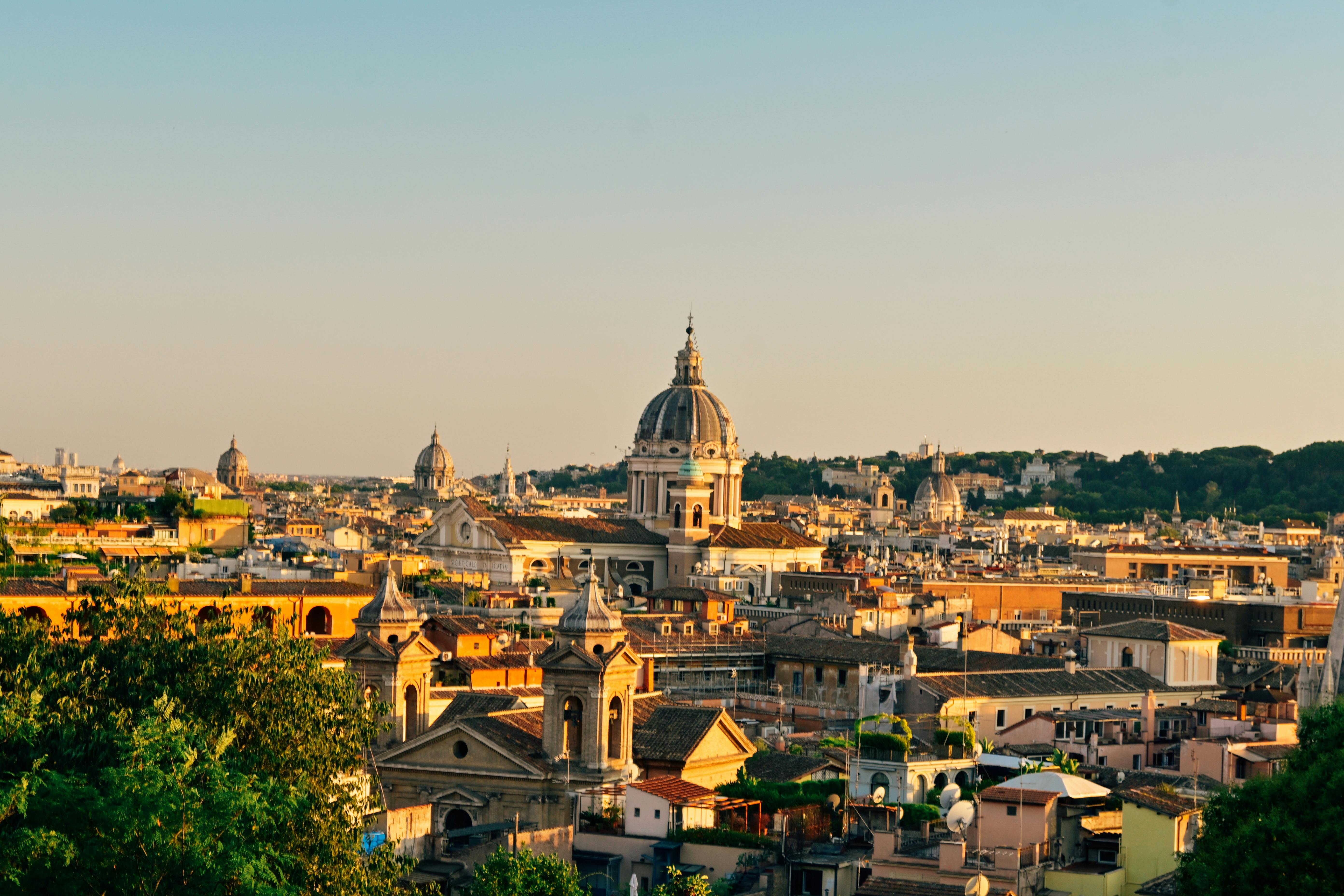 Panoramic aerial view of Rome skyline at sunset with historic terracotta rooftops, ancient domes, and iconic monuments glowing in warm golden light across the Eternal City