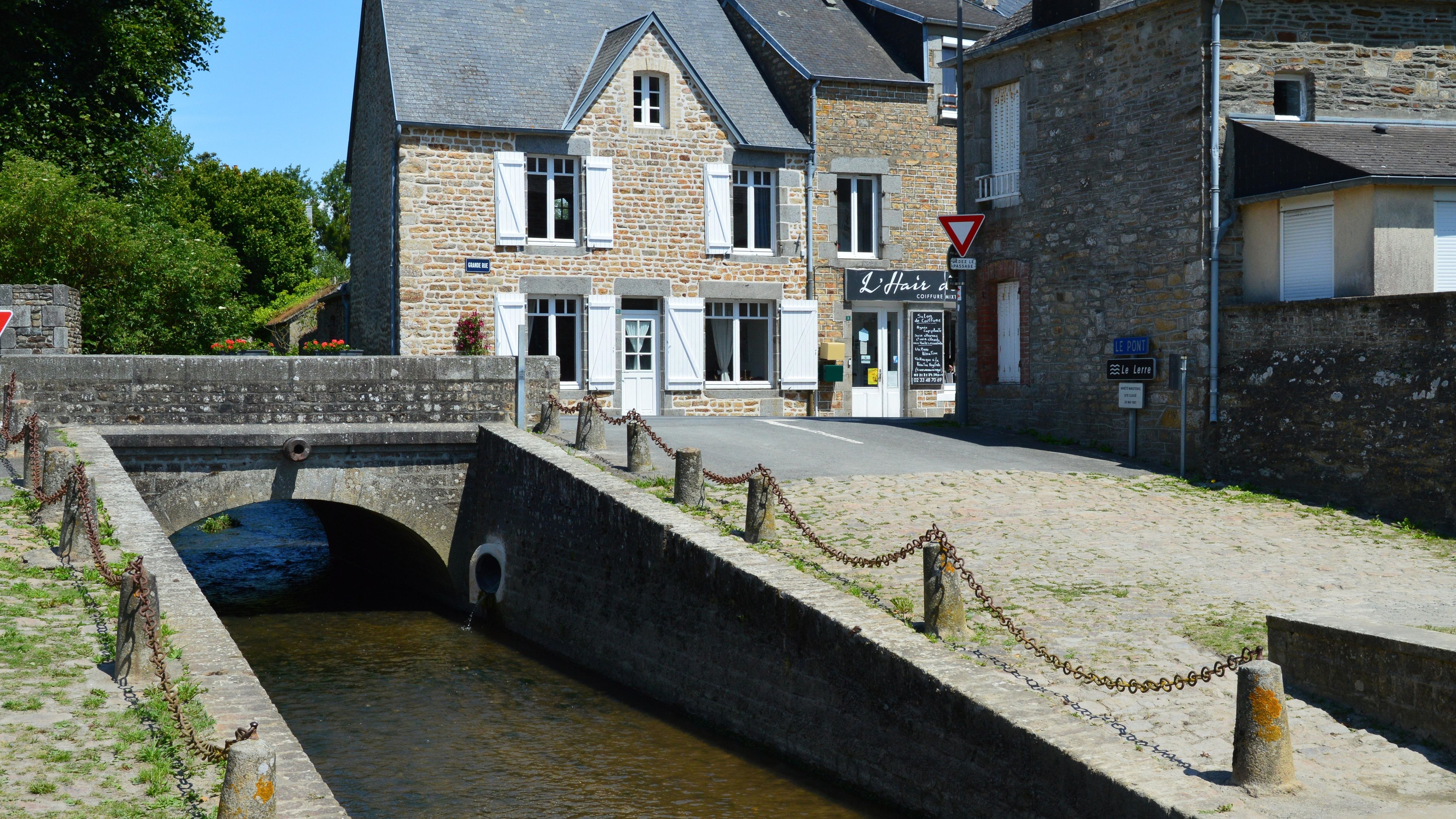 Picturesque stone house by a canal in Genêts, Normandie, France, under a clear blue sky.