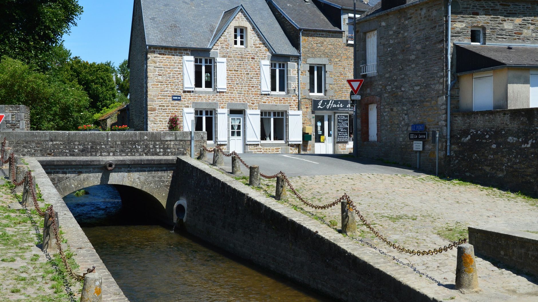 Picturesque stone house by a canal in Genêts, Normandie, France, under a clear blue sky.