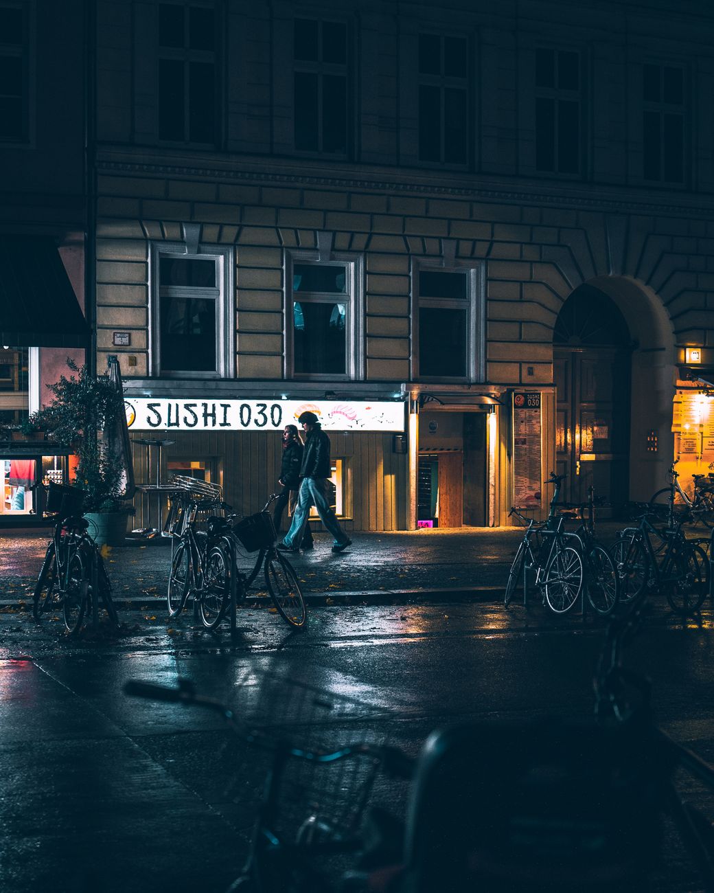 Moody urban nighttime scene on a wet Berlin street with bikes and a sushi shop.