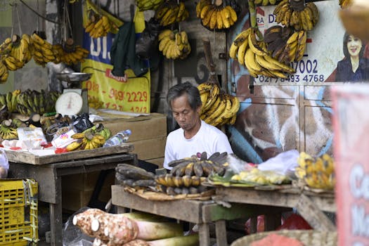 A street vendor sells bananas at an outdoor market, surrounded by hanging and stacked bananas.