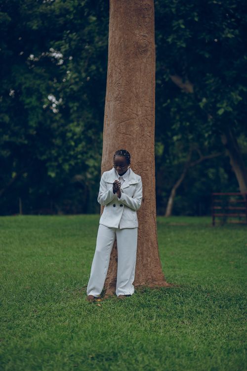 Free An individual in an elegant white suit stands contemplatively by a large tree in a lush green park. Stock Photo
