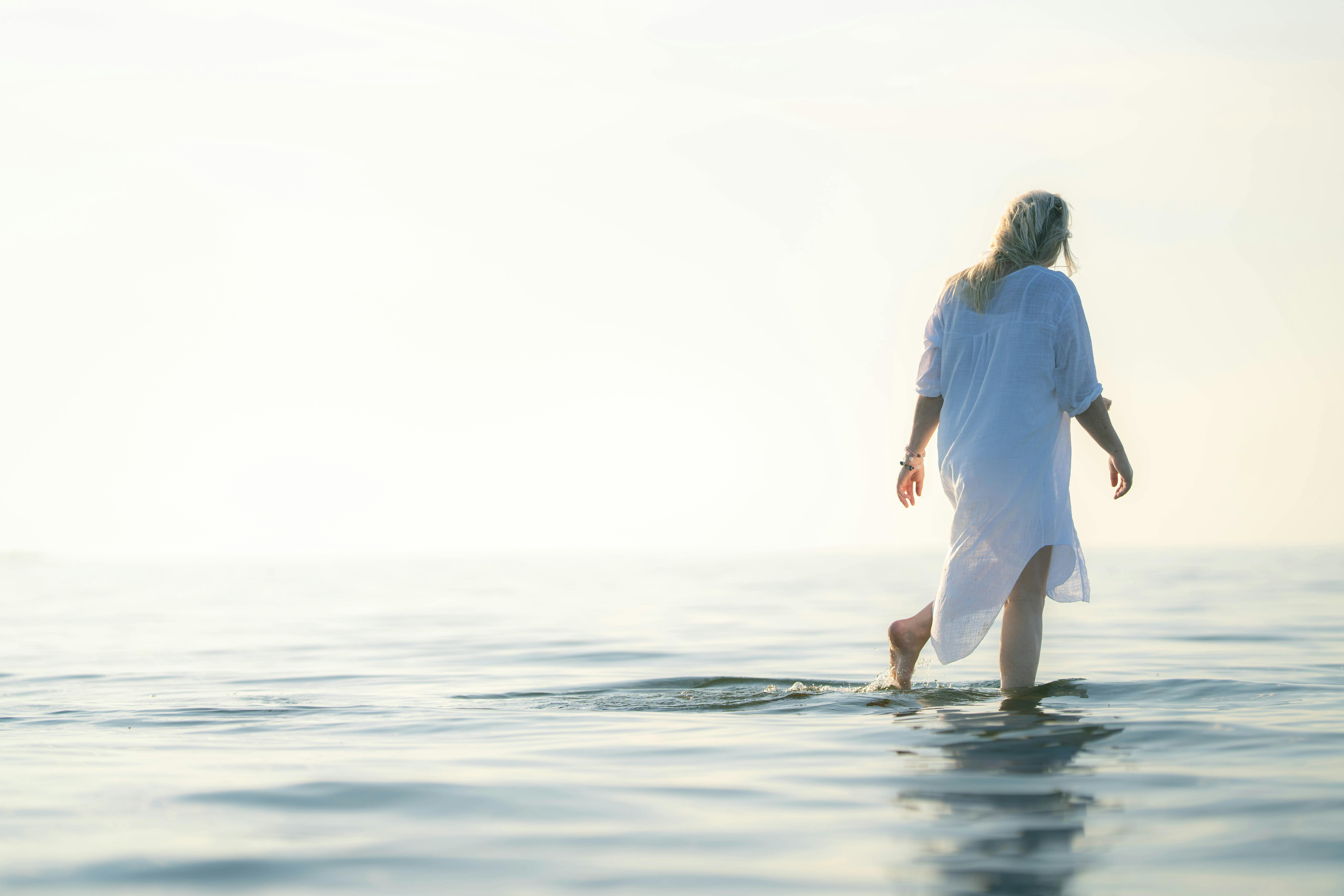 Une femme vêtue d'une robe blanche marche dans les eaux tranquilles de l'océan au lever du soleil, créant une scène paisible et réfléchie.