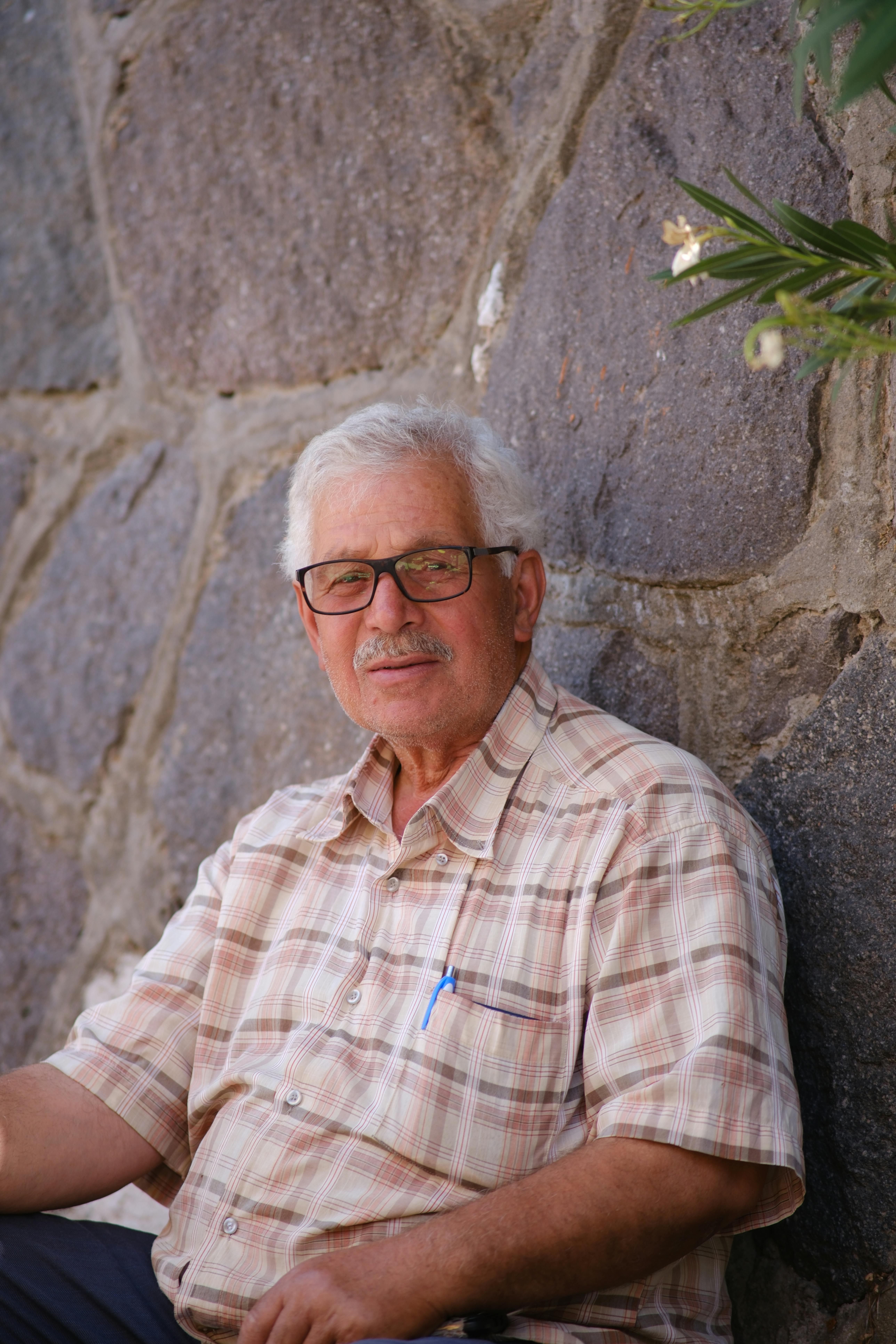 Portrait of an elderly man sitting against a stone wall outdoors in İzmir, Türkiye.
