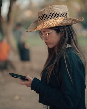 A woman wearing a straw hat and glasses texting in an outdoor park in Buenos Aires, Argentina.