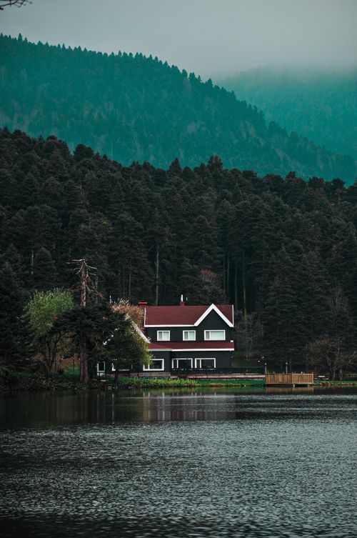 Free Beautiful red-roofed house by a serene lakeside surrounded by dense forest and mountains. Stock Photo