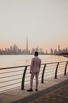 一位穿着正式服装的时尚男士，正站在迪拜海滨欣赏日落时的城市天际线