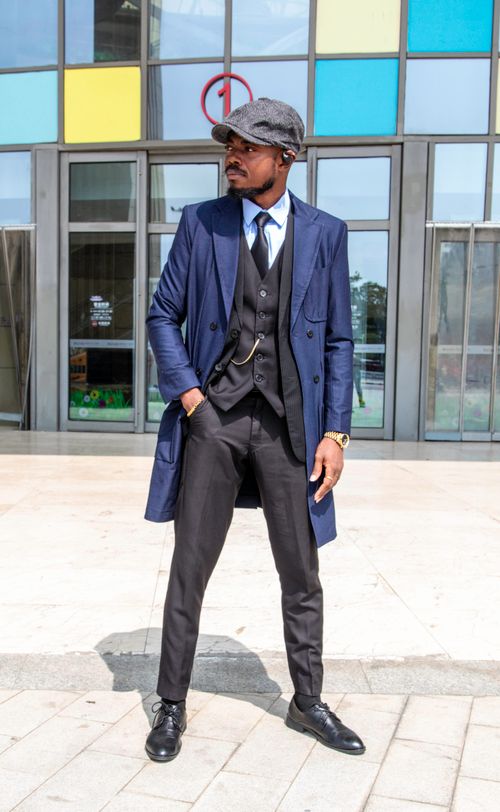 Free Man in a blue vintage suit and flat cap, posing outside modern building. Fashionable and confident. Stock Photo