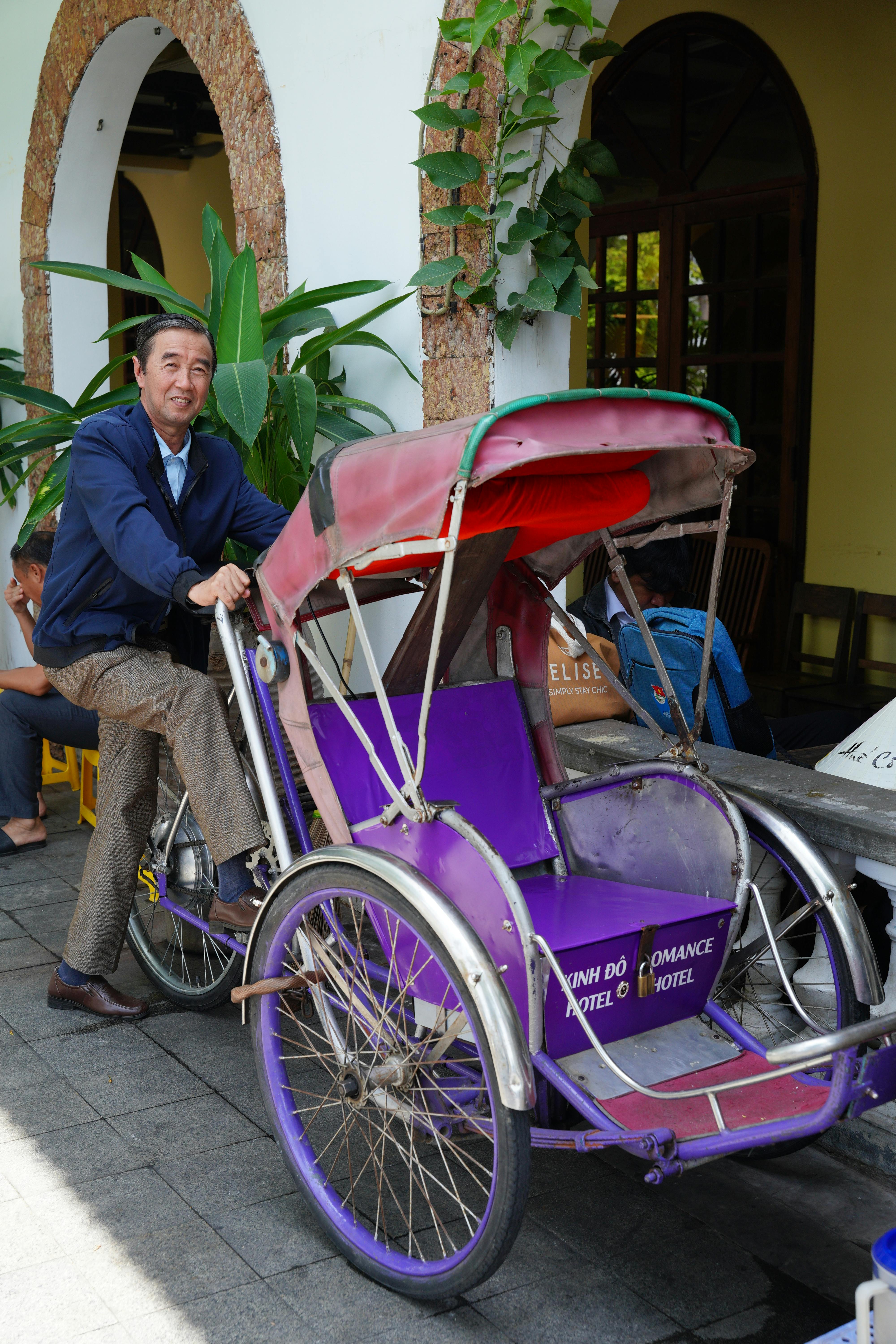 Free A man rides a vibrant cyclo by a Vietnamese hotel entrance, showcasing local transportation. Stock Photo